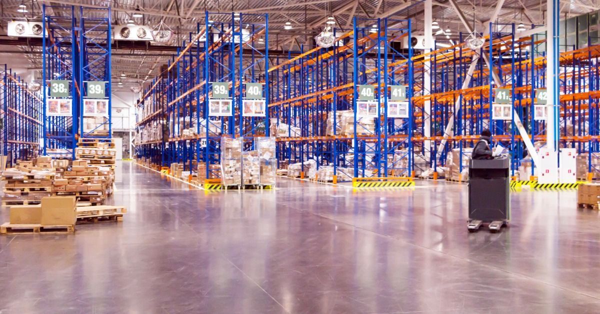 A large warehouse aisle with tall blue and orange pallet racks, stacked boxes, and overhead lights.