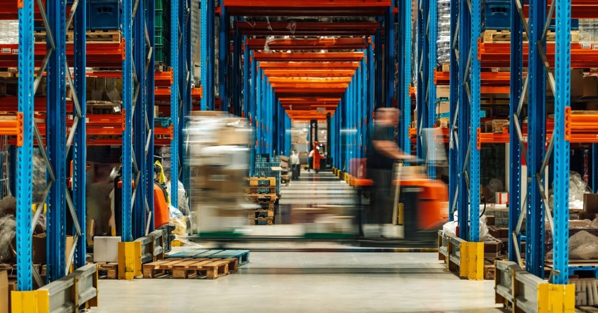 A warehouse aisle lined with blue and orange racks, with blurred forklifts moving pallets along the floor.