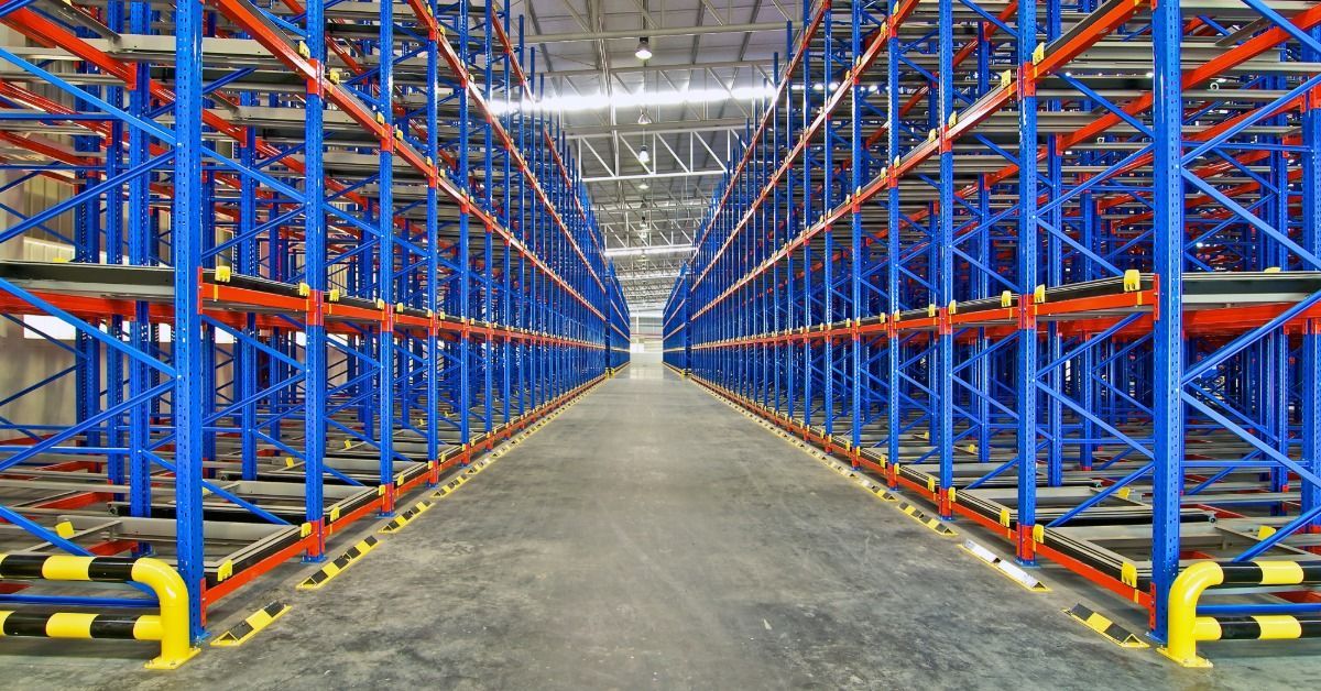 An empty warehouse aisle between tall blue and red metal storage racks, with overhead lights and a concrete floor.