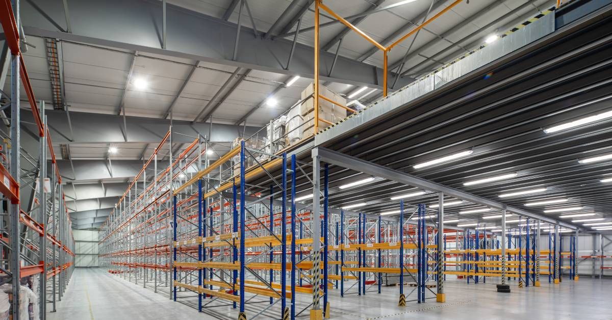 The interior of a well-lit, empty warehouse. There is a mezzanine built next to multiple pallet racks.