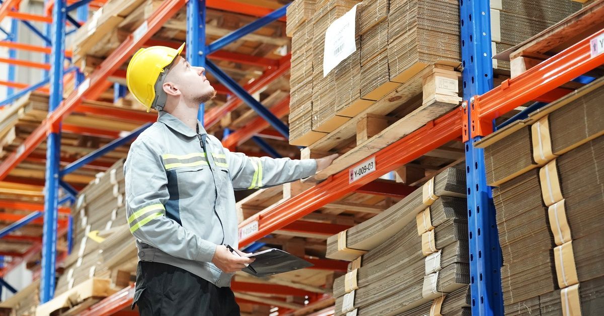 A warehouse worker checking the inventory stacked on a pallet racking shelf. He has a clipboard in his hand.