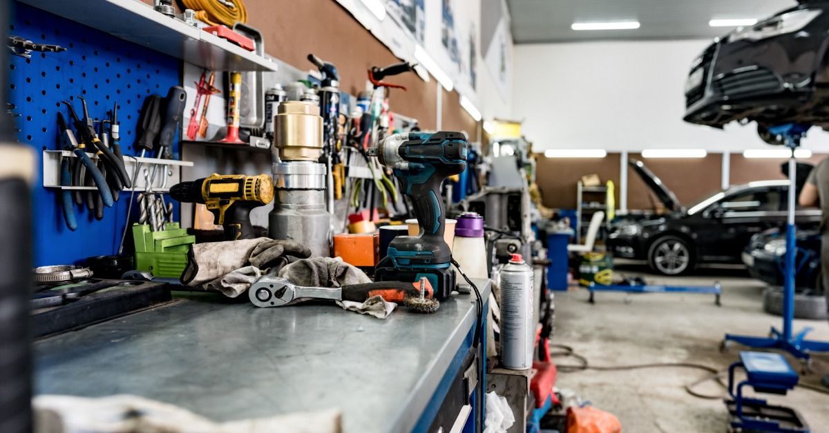 A large workbench in an automotive repair shop filled with tools. Behind the benches are two cars awaiting repair.