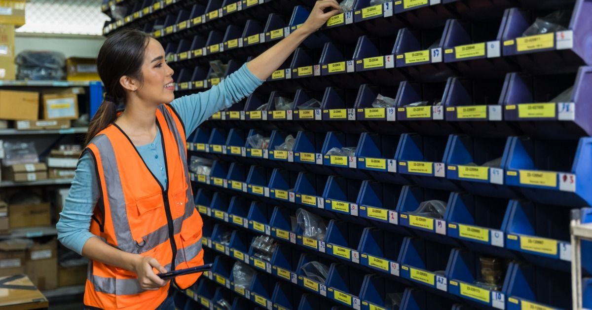 A woman in an orange safety vest holding a tablet and reaching toward labeled blue bins on warehouse shelves.