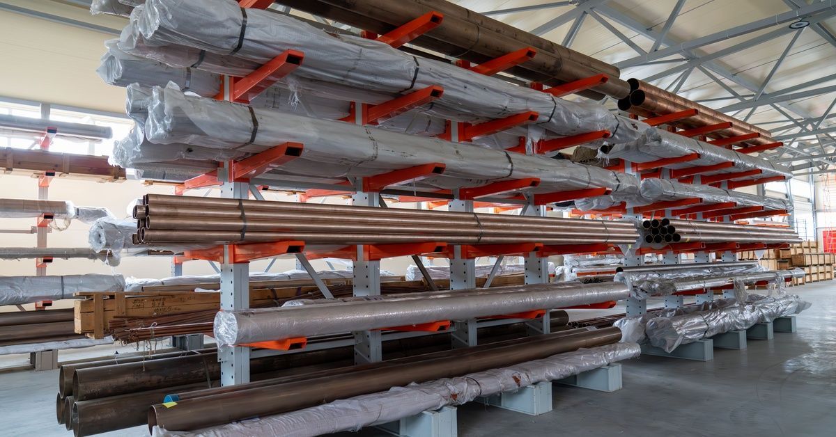 A warehouse rack with red arms holding long metal pipes, wrapped bundles, and stacked tubes under a