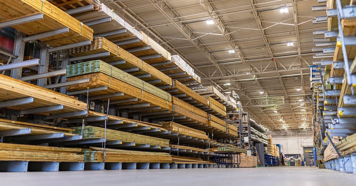 A warehouse aisle with stacked lumber boards stored on tall orange cantilever racks under bright ceiling lights.