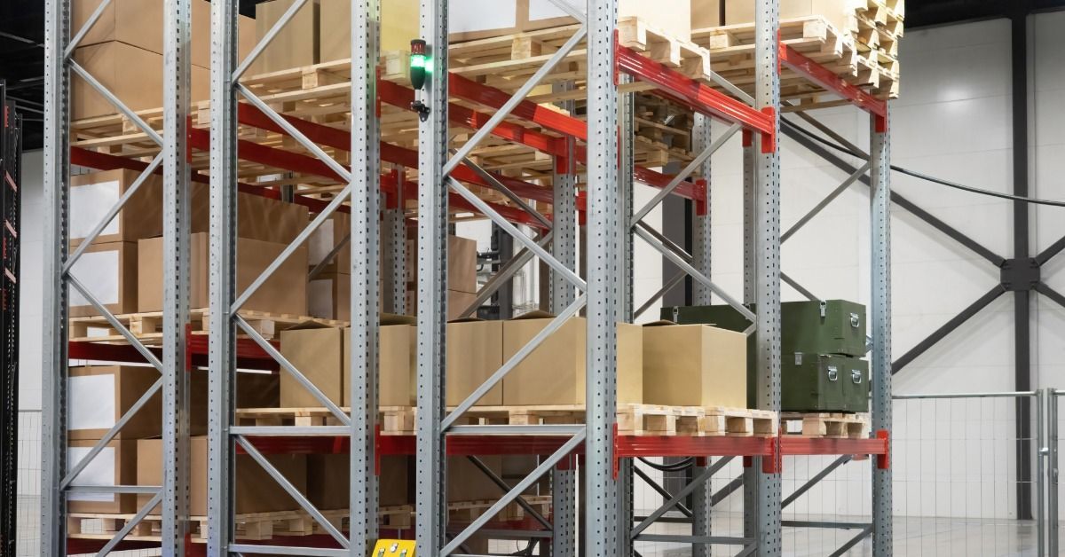 A warehouse aisle with boxes stacked on pallets and tall metal shelving under bright ceiling lights.