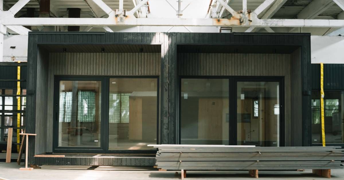 A black, modern-style modular prefabricated structure set up inside a warehouse. The front wall has two large sliding glass doors.
