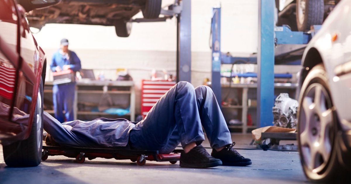 A mechanic is working in an auto repair shop. He lays on his back as he tinkers underneath a parked car.