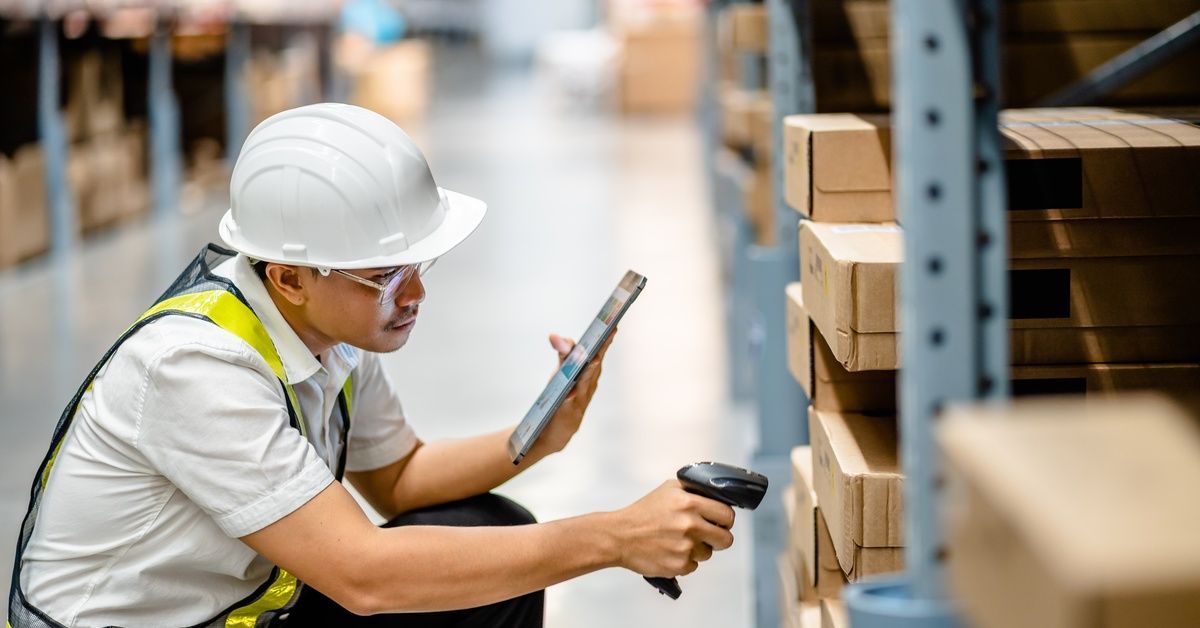 A male warehouse worker in a hard hat using a handheld scanner and a digital tablet to check warehouse inventory.
