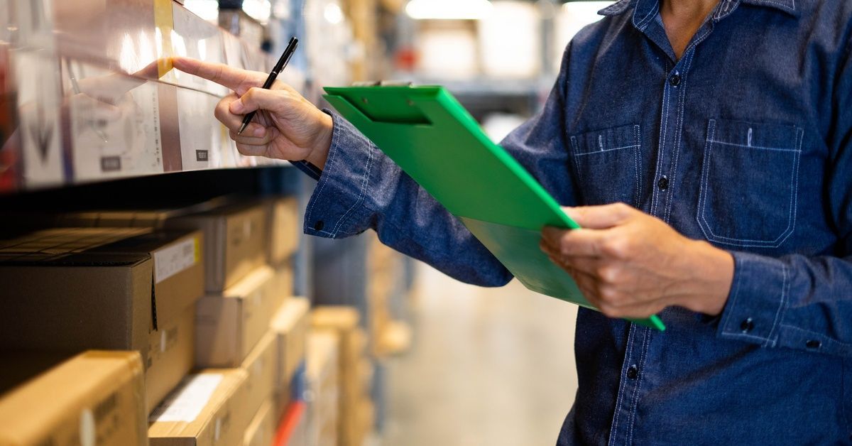 A warehouse worker holding a clipboard in one hand while pointing at a label on an industrial shelving unit with the other.