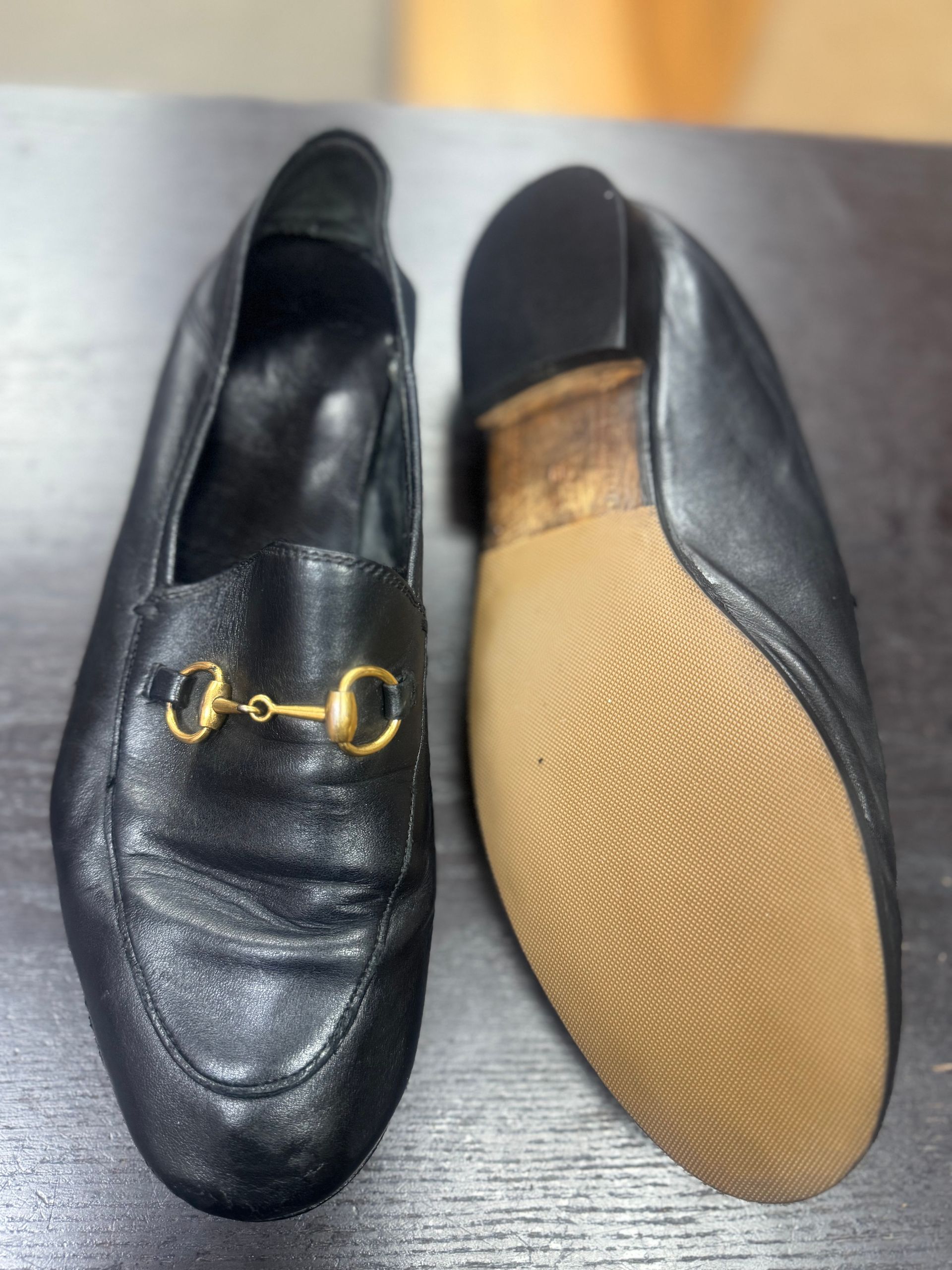 A pair of black leather loafers with gold-toned horsebit hardware, shown from the top and the beige rubber sole.