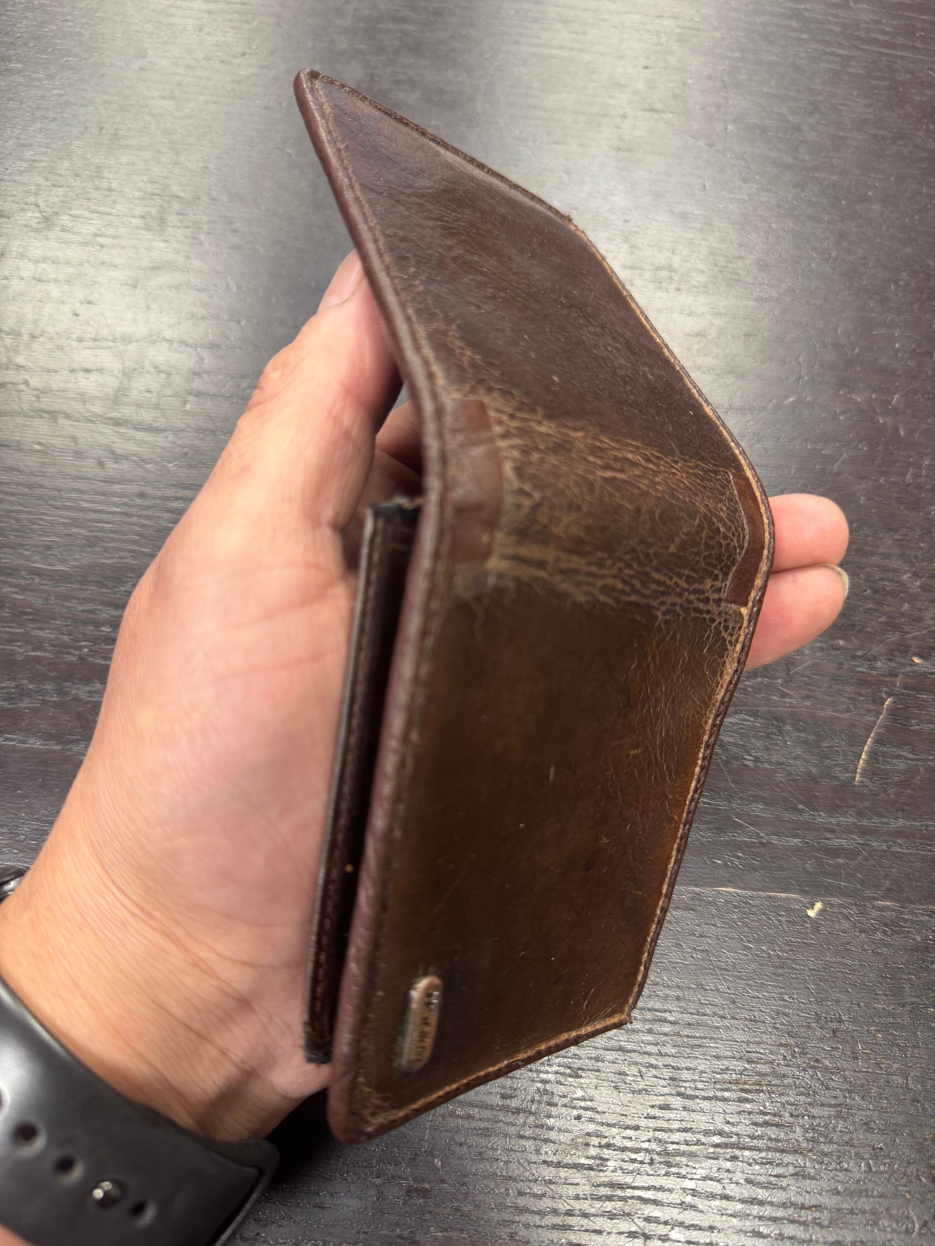 A person holds a worn, folded brown leather wallet with a small metal logo on a dark tabletop surface.