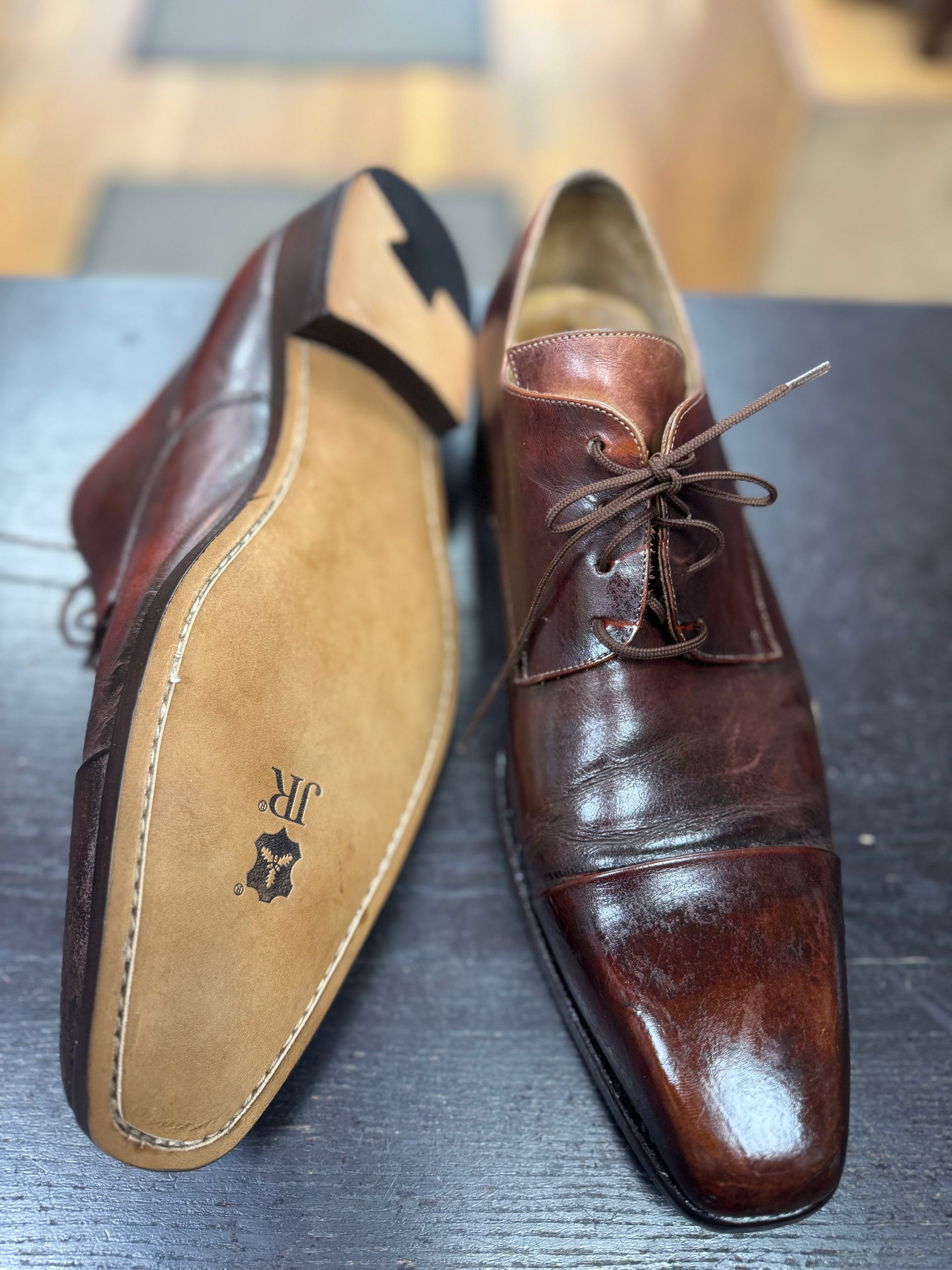 Two brown leather oxford shoes on a dark surface, one showing its smooth, branded JR leather sole and the other its toe.