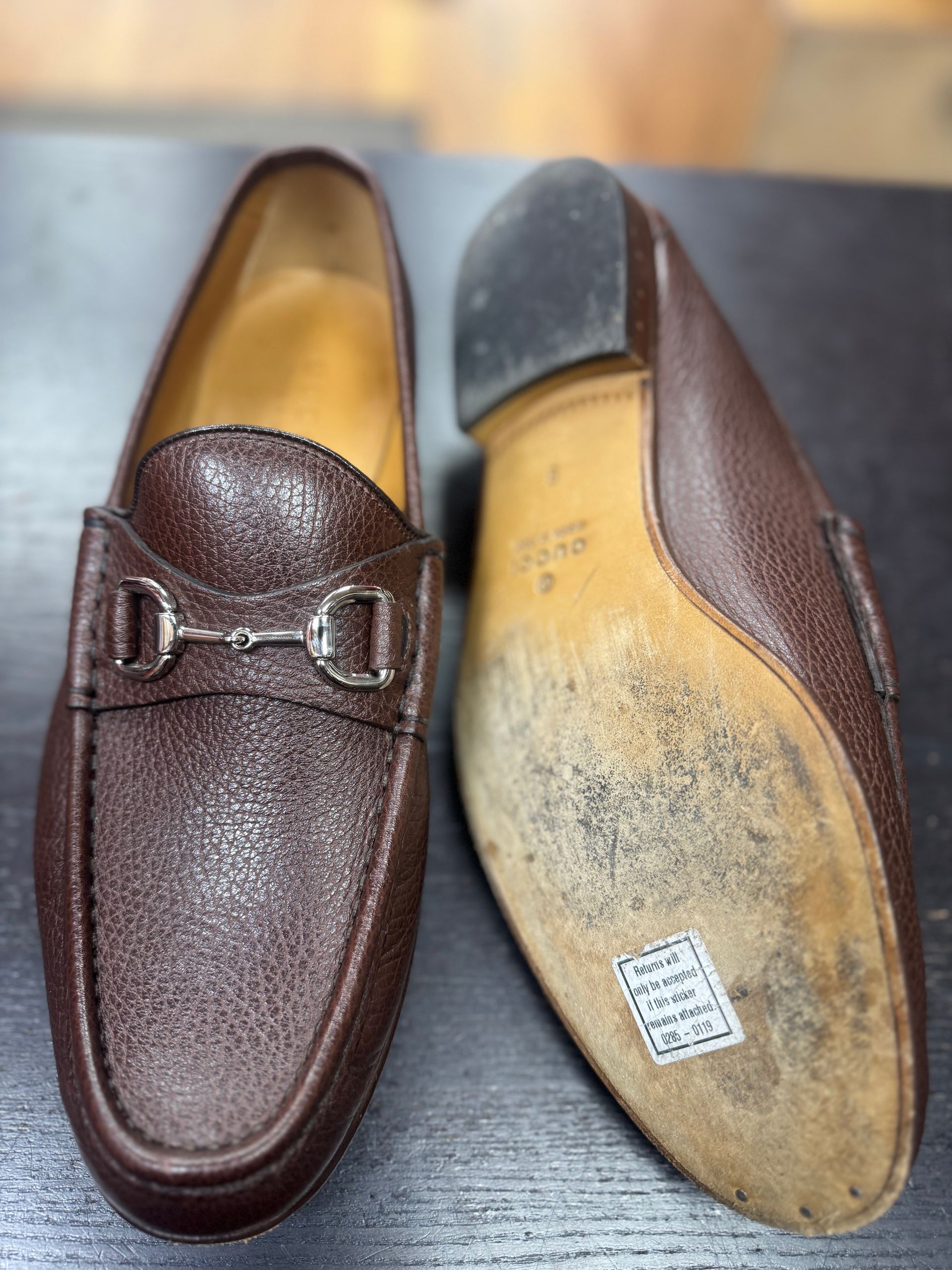 A pair of brown pebbled leather loafers with silver horsebit hardware, one shown from the top and one from the sole.
