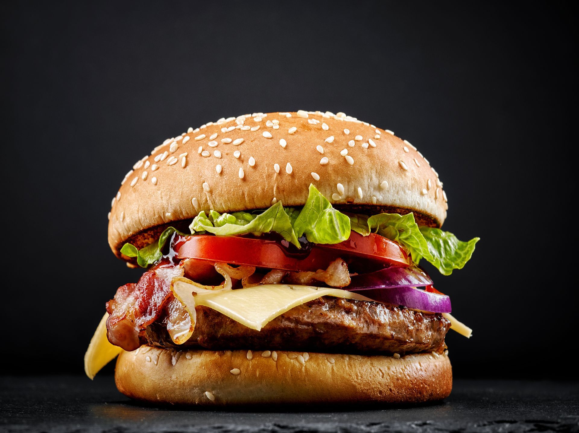 Burger mit Sesambrötchen, Salat, Tomaten, Zwiebeln, Speck und Käse auf dunklem Untergrund.