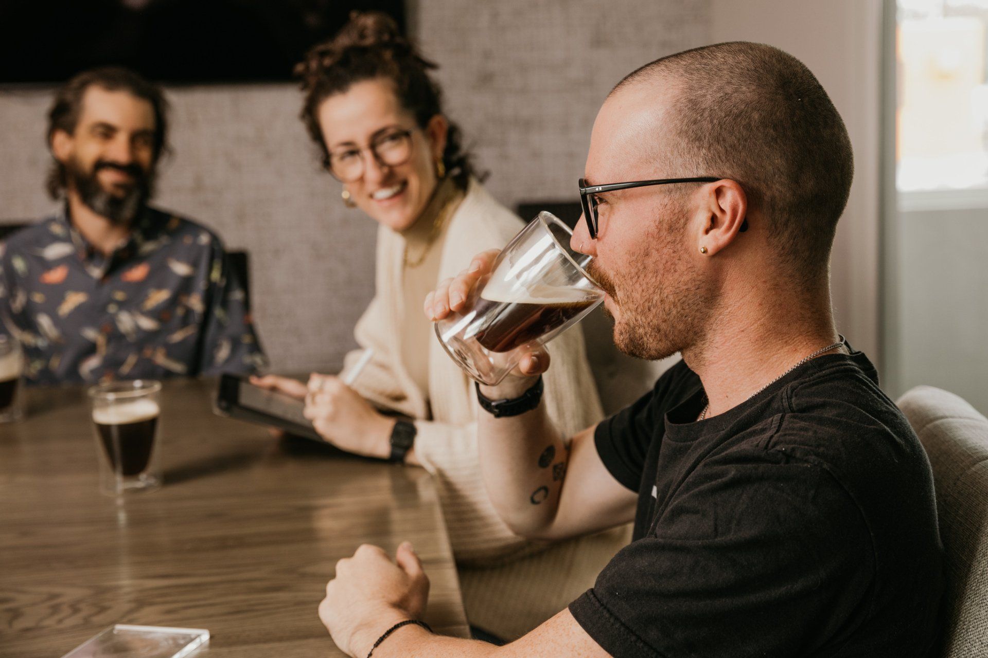 Man drinks dark beer at a table with two smiling people.