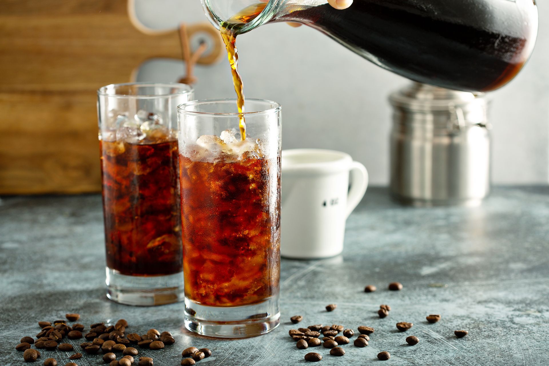 Two tall glasses filled with ice and coffee are being poured, surrounded by coffee beans.