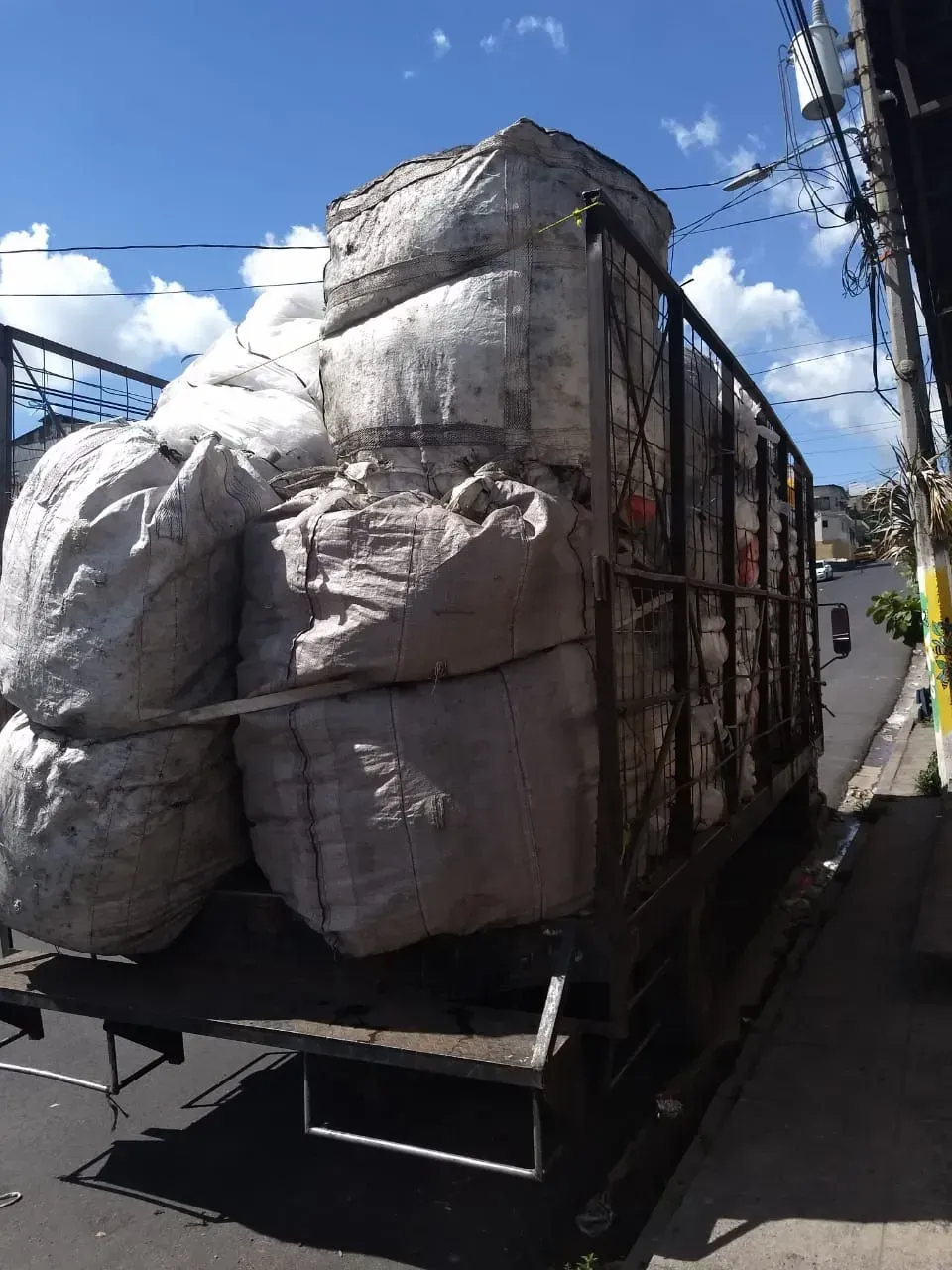 Un camión grande cargado con voluminosos sacos grises circula por una calle estrecha bajo un cielo azul.