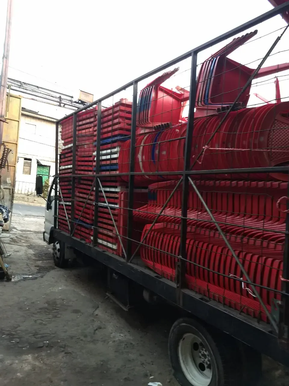Camión cargado con sillas metálicas rojas apiladas en un estrecho callejón al aire libre.