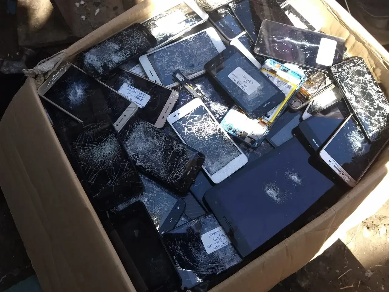 Caja de cartón llena de teléfonos inteligentes variados y cristales rotos bajo la luz del sol.
