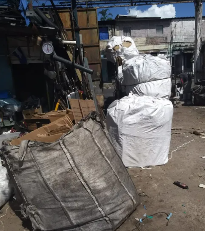 Grandes sacos industriales blancos apilados en un patio de trabajo al aire libre, con una bolsa gris en primer plano.