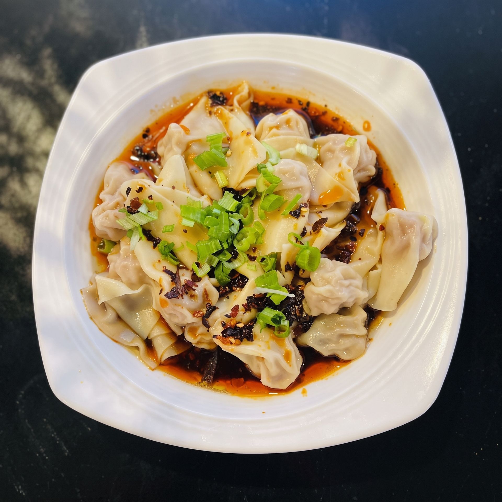 Steamed dumplings in spicy red chili oil, topped with chopped green herbs, in a white bowl.