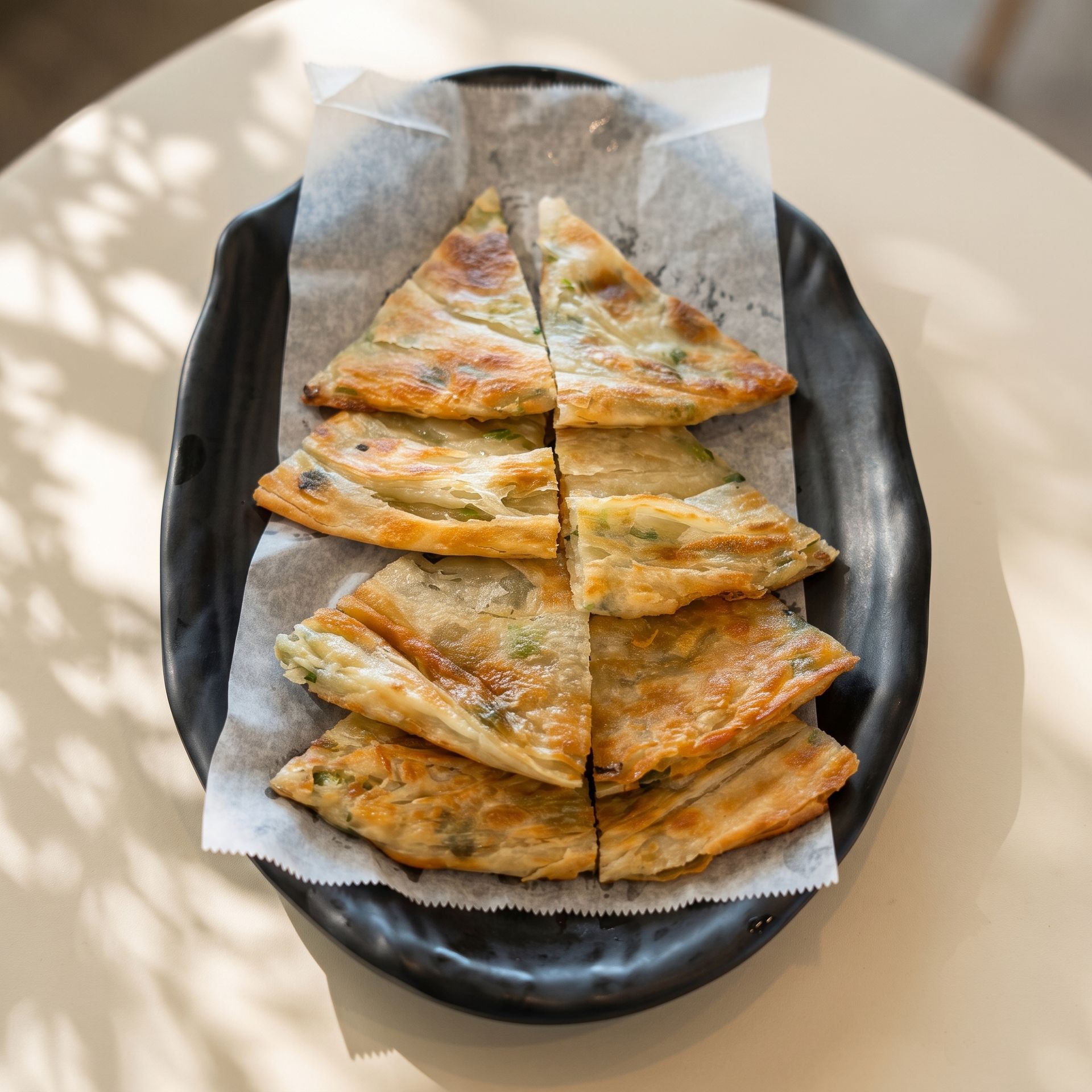 Sliced golden flatbread on parchment in a black oval dish, lit by soft sunlight.