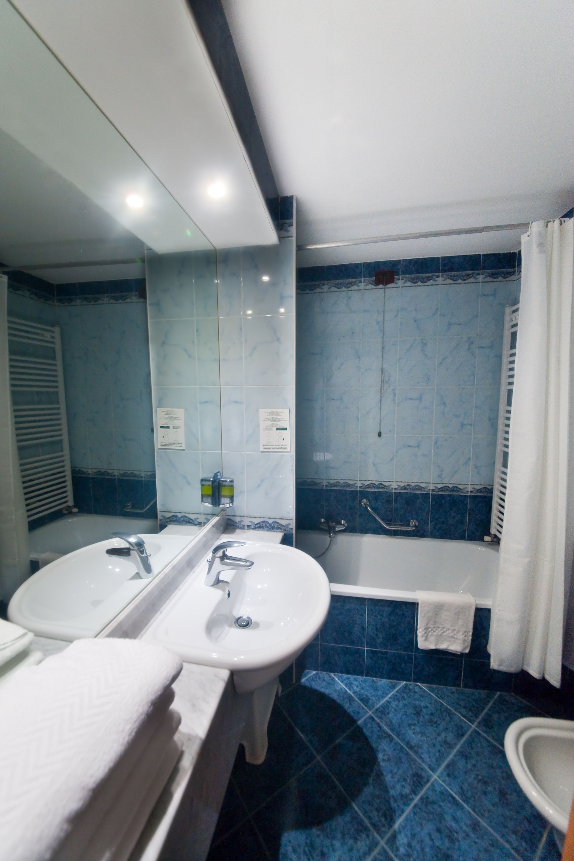 Bagno con pareti piastrellate blu, lavandino bianco, vasca e asciugamani; grande specchio.