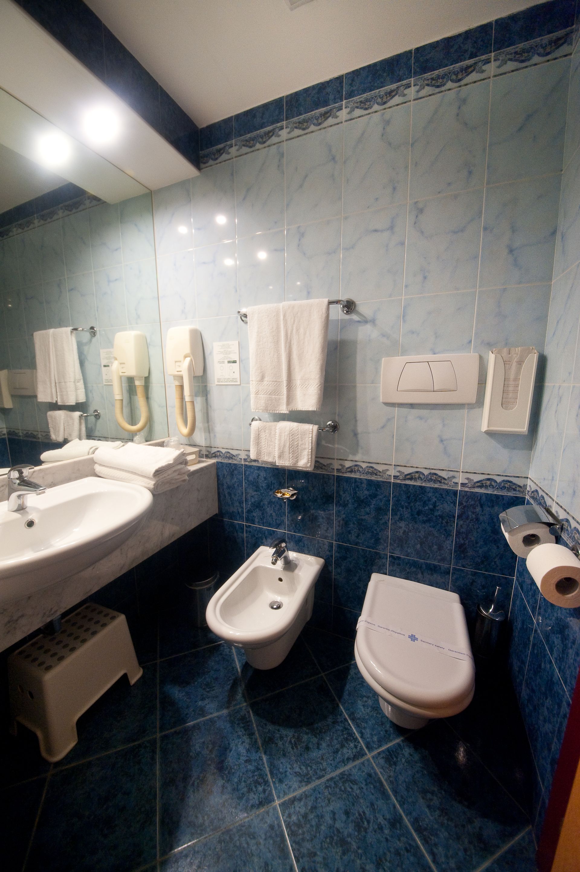 Bagno con piastrelle blu, WC, bidet, lavandino, asciugamani e sgabello.