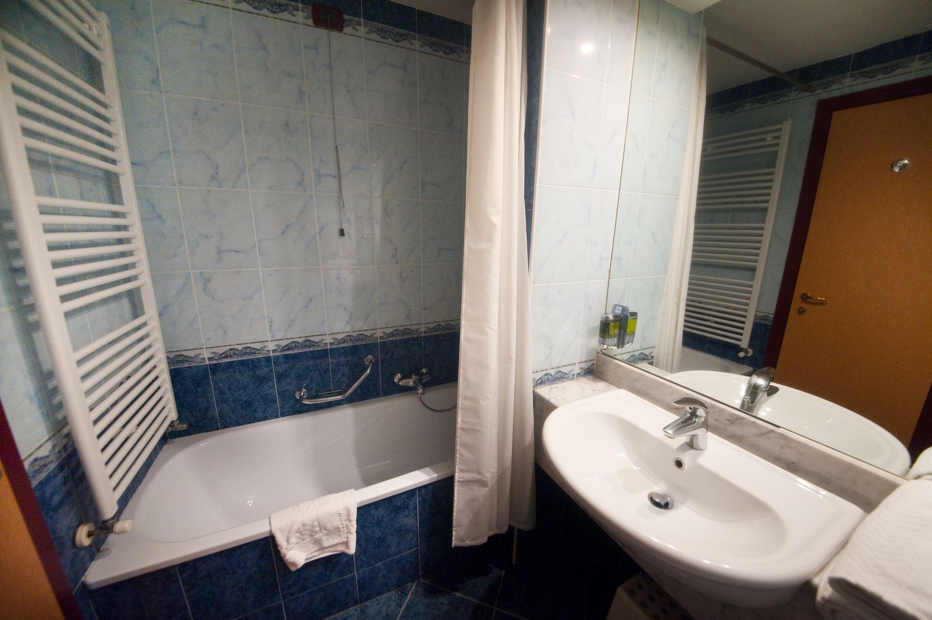 Bagno con vasca da bagno bianca, lavandino e termosifone su pareti piastrellate di blu. Tenda da doccia appesa.