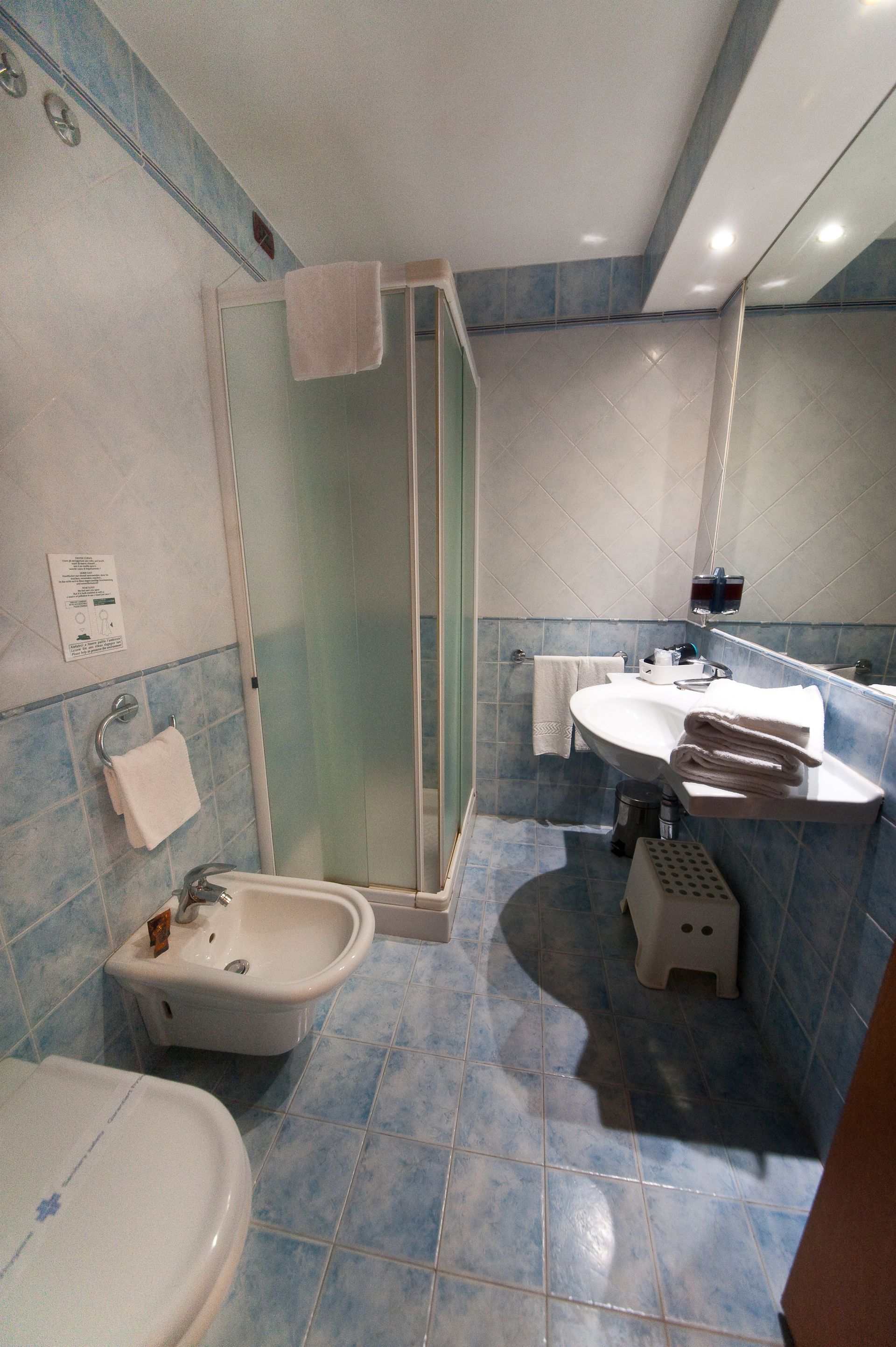 Bagno con piastrelle blu, doccia, WC, bidet, lavandino, specchio e un piccolo sgabello.