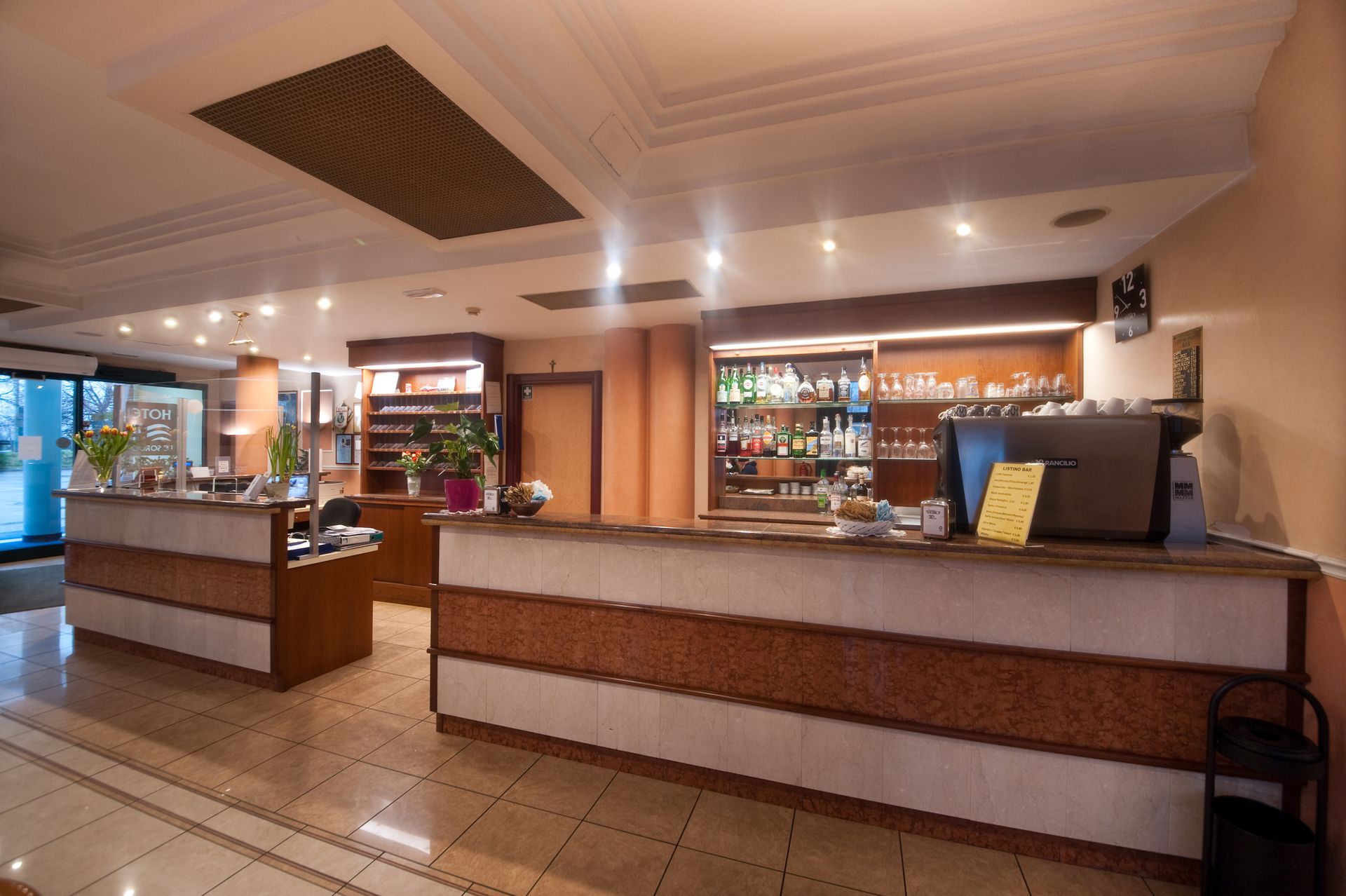 Reception dell'hotel con due banconi e un bar. Toni beige e marrone.