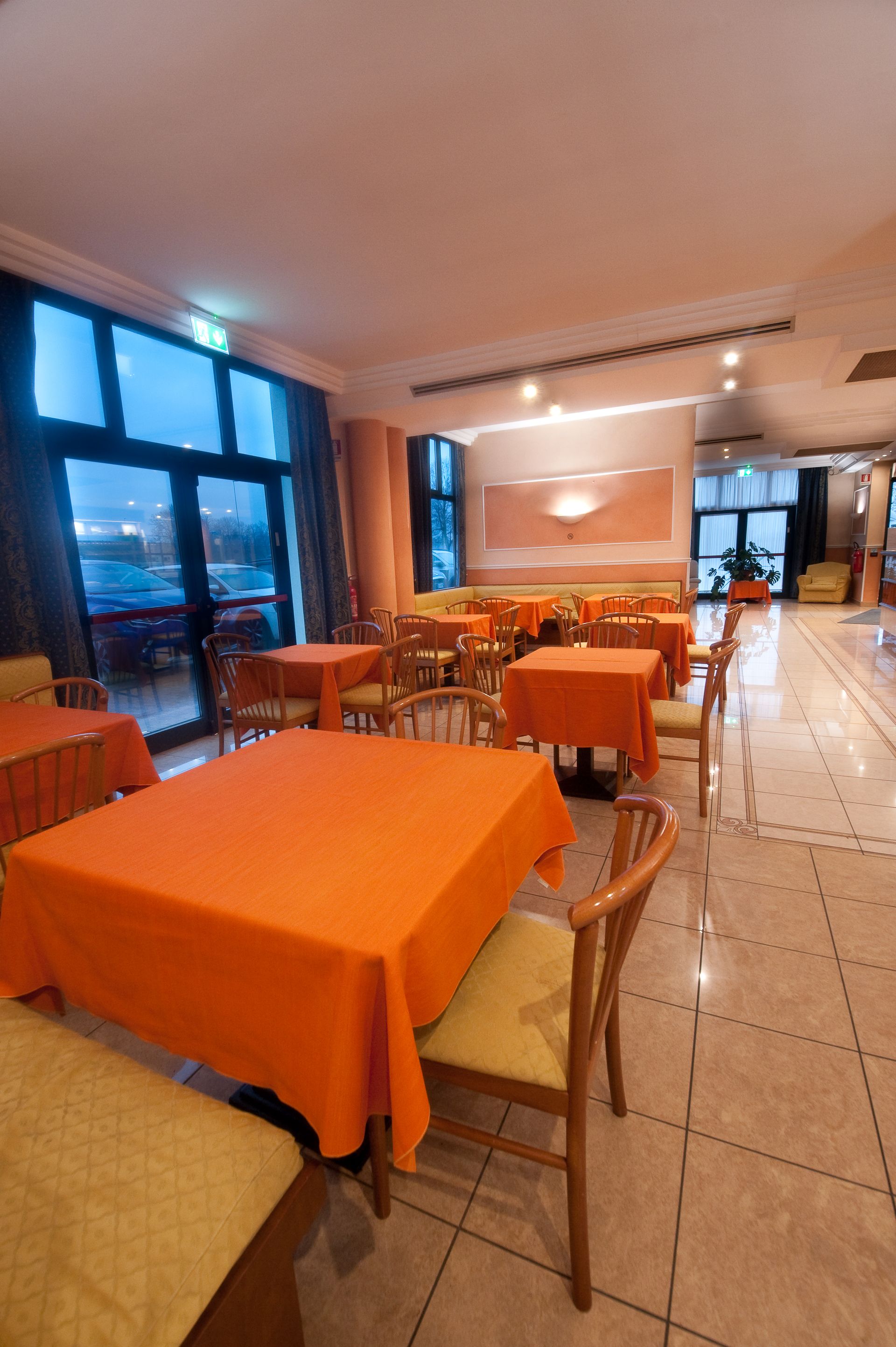 Interno del ristorante con tavoli quadrati coperti da tovaglie arancioni e sedie in legno. Ampia finestra.