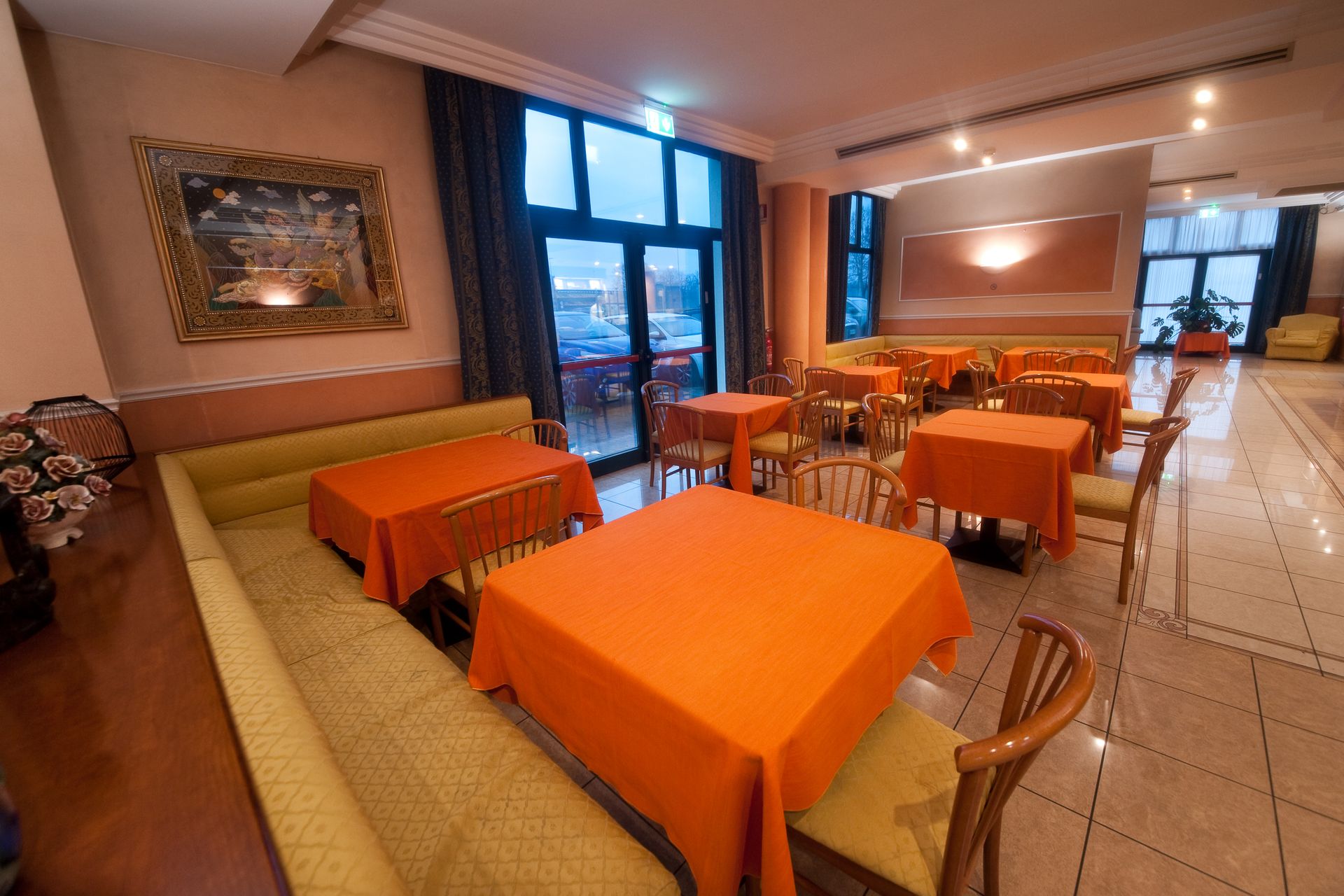 Interno del ristorante con tovaglie e tende arancioni, luce naturale e un dipinto.