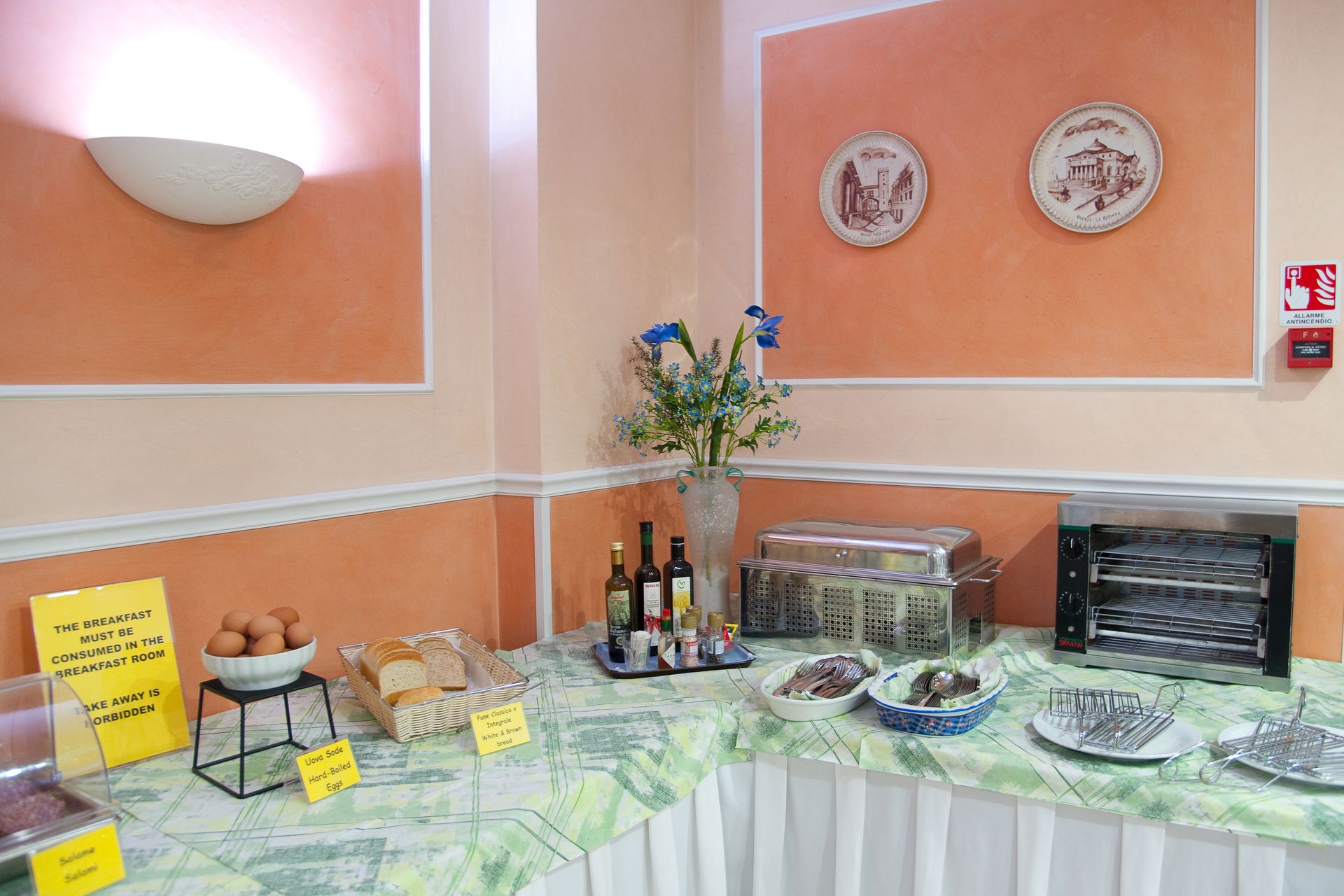 Colazione a buffet con cibo e scaldavivande su un tavolo, uova, pane e decorazioni contro una parete arancione.