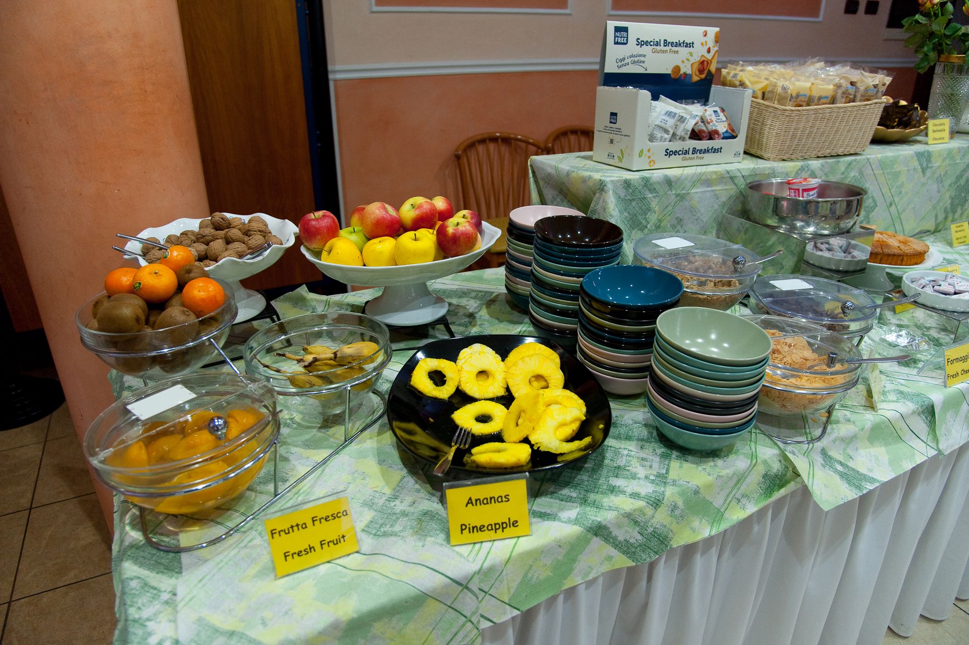 Tavolo da buffet con frutta varia: mele, arance, ananas, kiwi. Ciotole e piatti da portata su una tovaglia floreale.