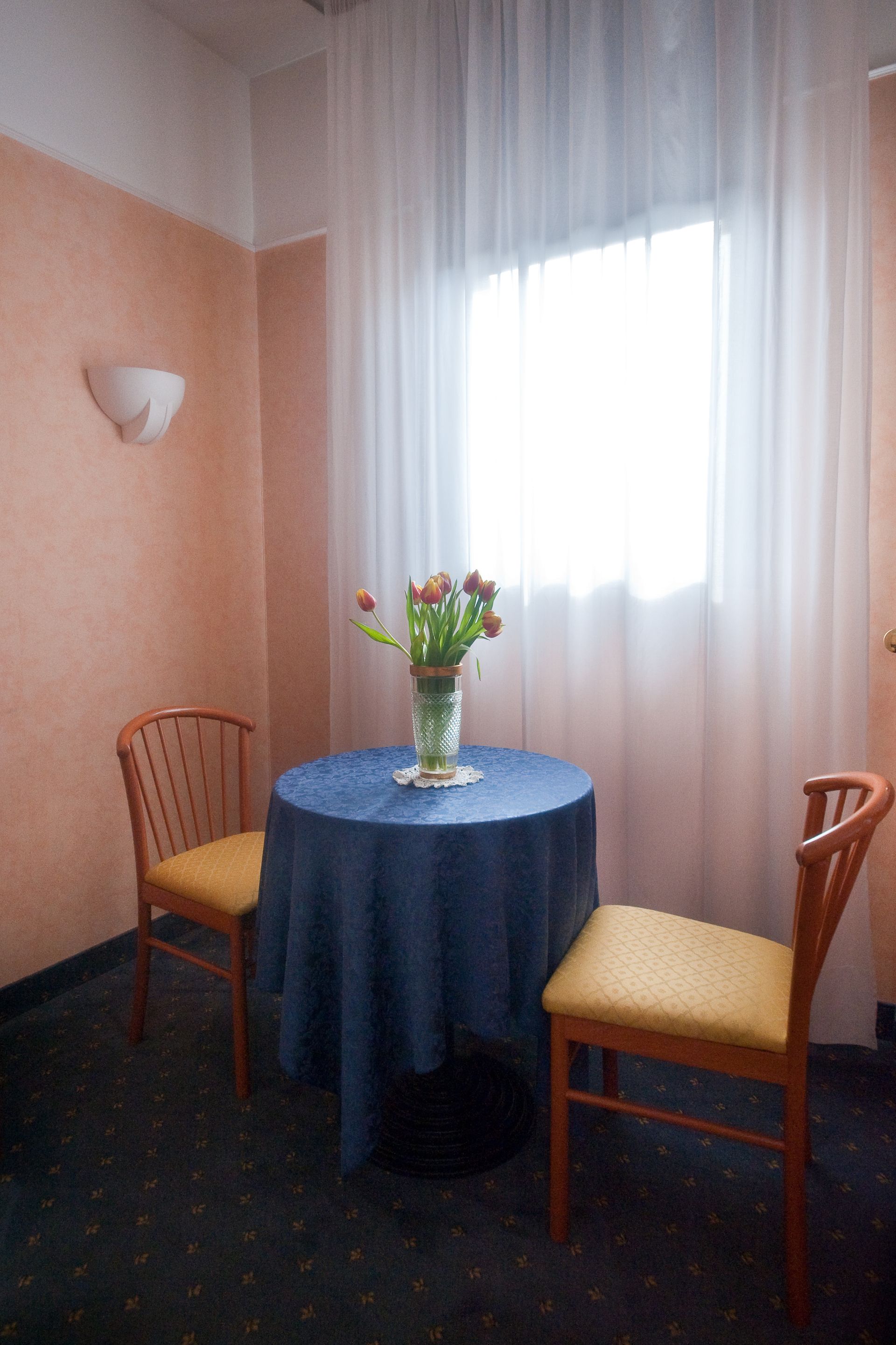 Tavolino con tovaglia blu, due sedie e un vaso di fiori vicino a una finestra con tende trasparenti.