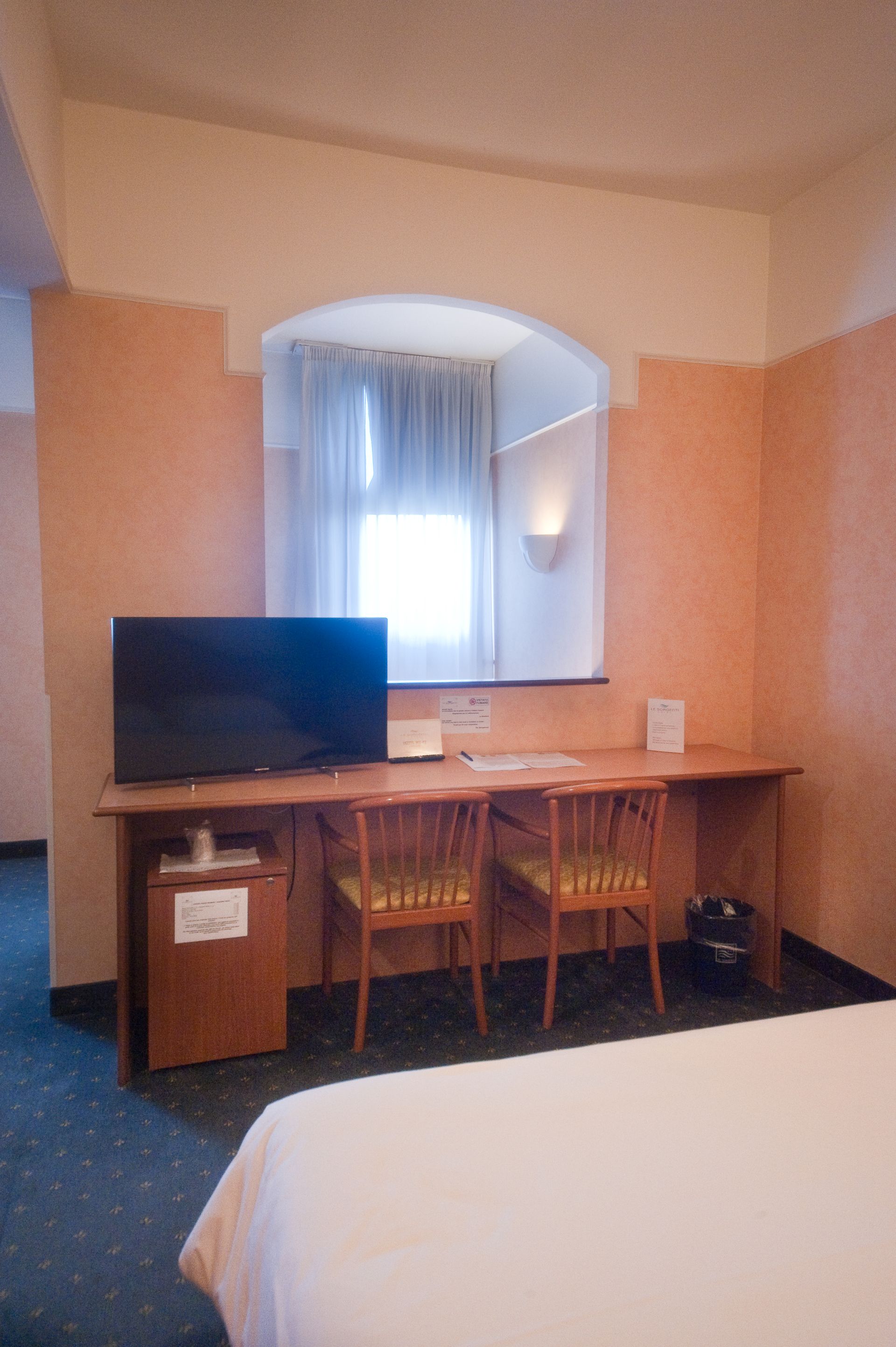 Interno di una camera d'albergo con scrivania, due sedie, TV e finestra con tende. Pareti color pesca.