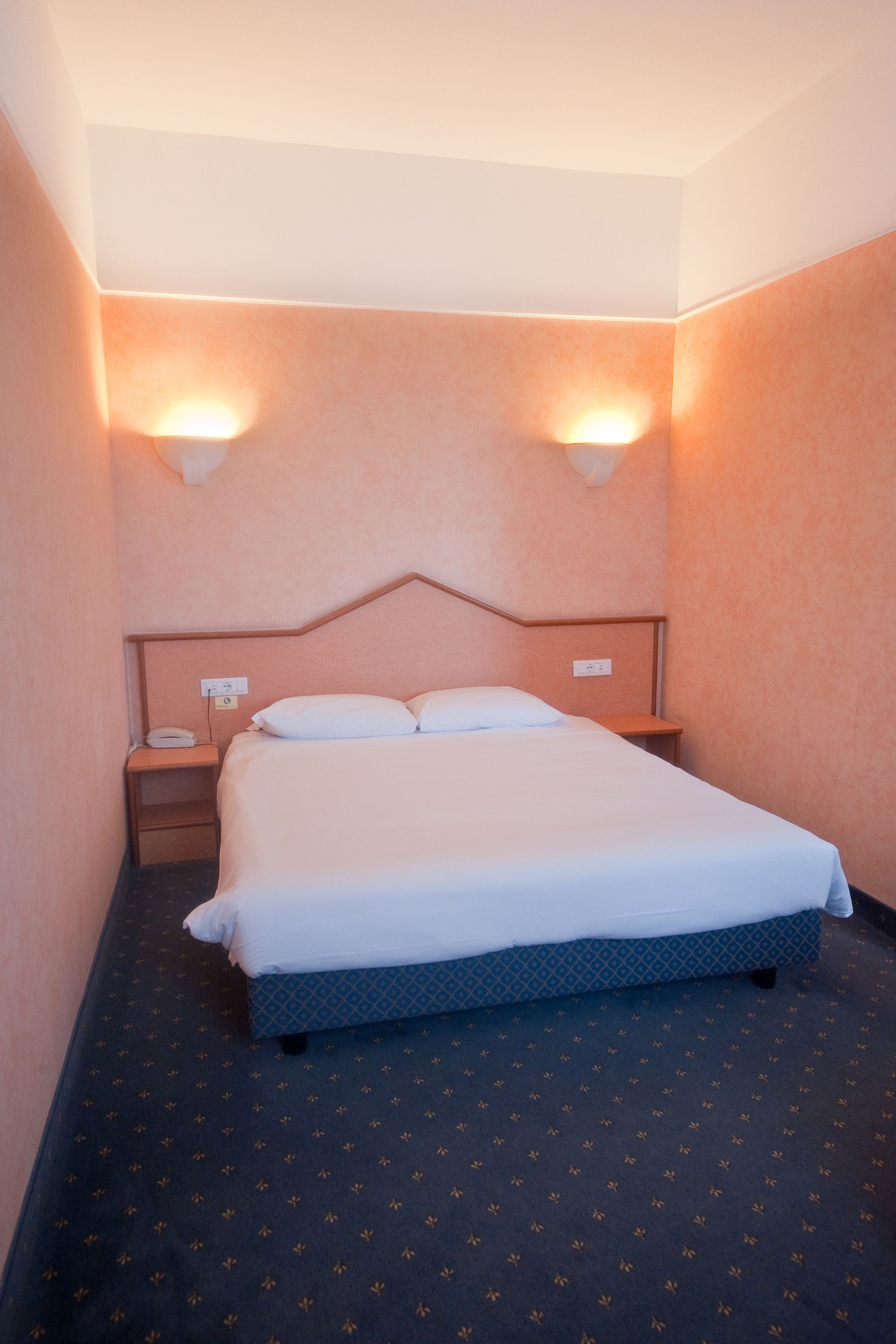Camera d'albergo con letto matrimoniale, pareti color pesca, due comodini, moquette blu e due applique.