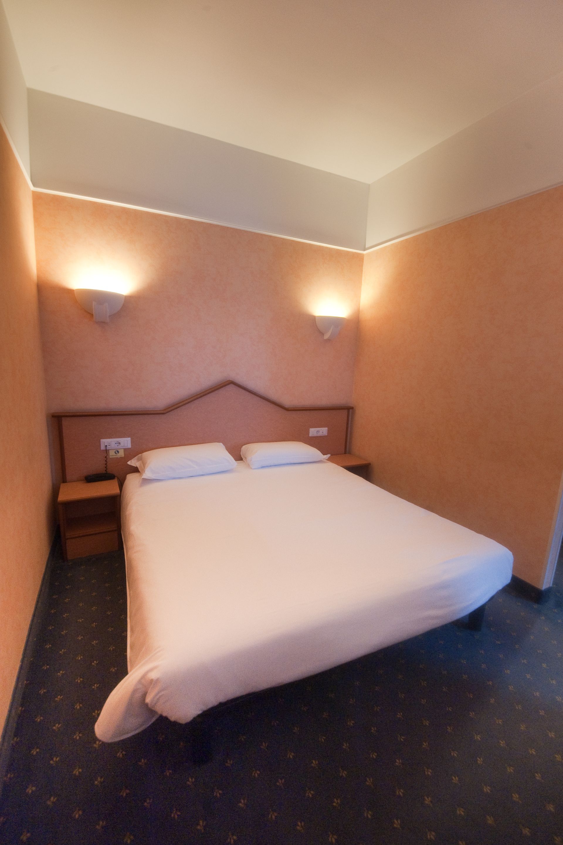 Una camera d'albergo con un letto matrimoniale, due comodini e applique su pareti color pesca.