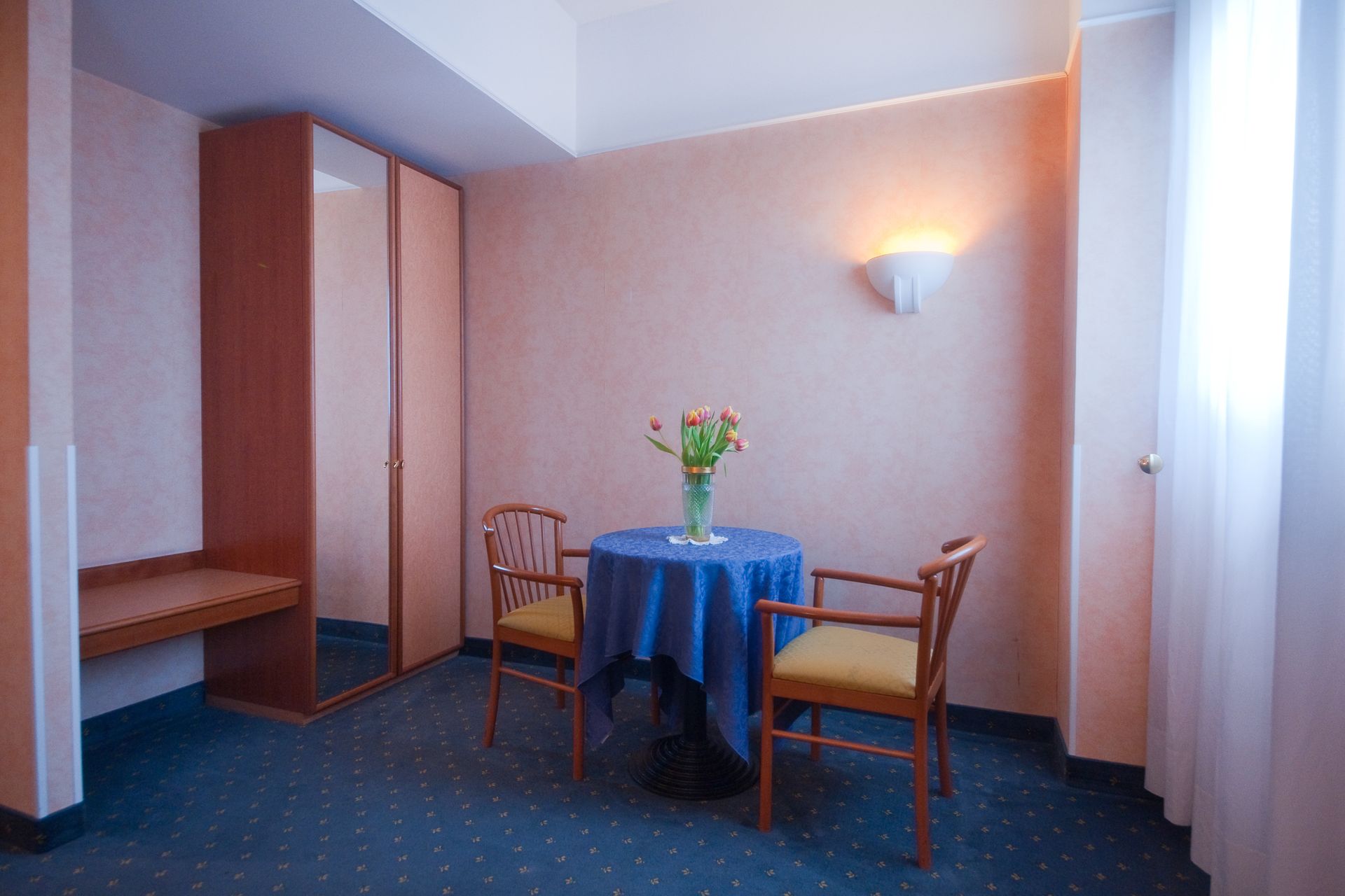 Camera d'albergo con tavolo e sedie, armadio, scrivania e finestra con tende.