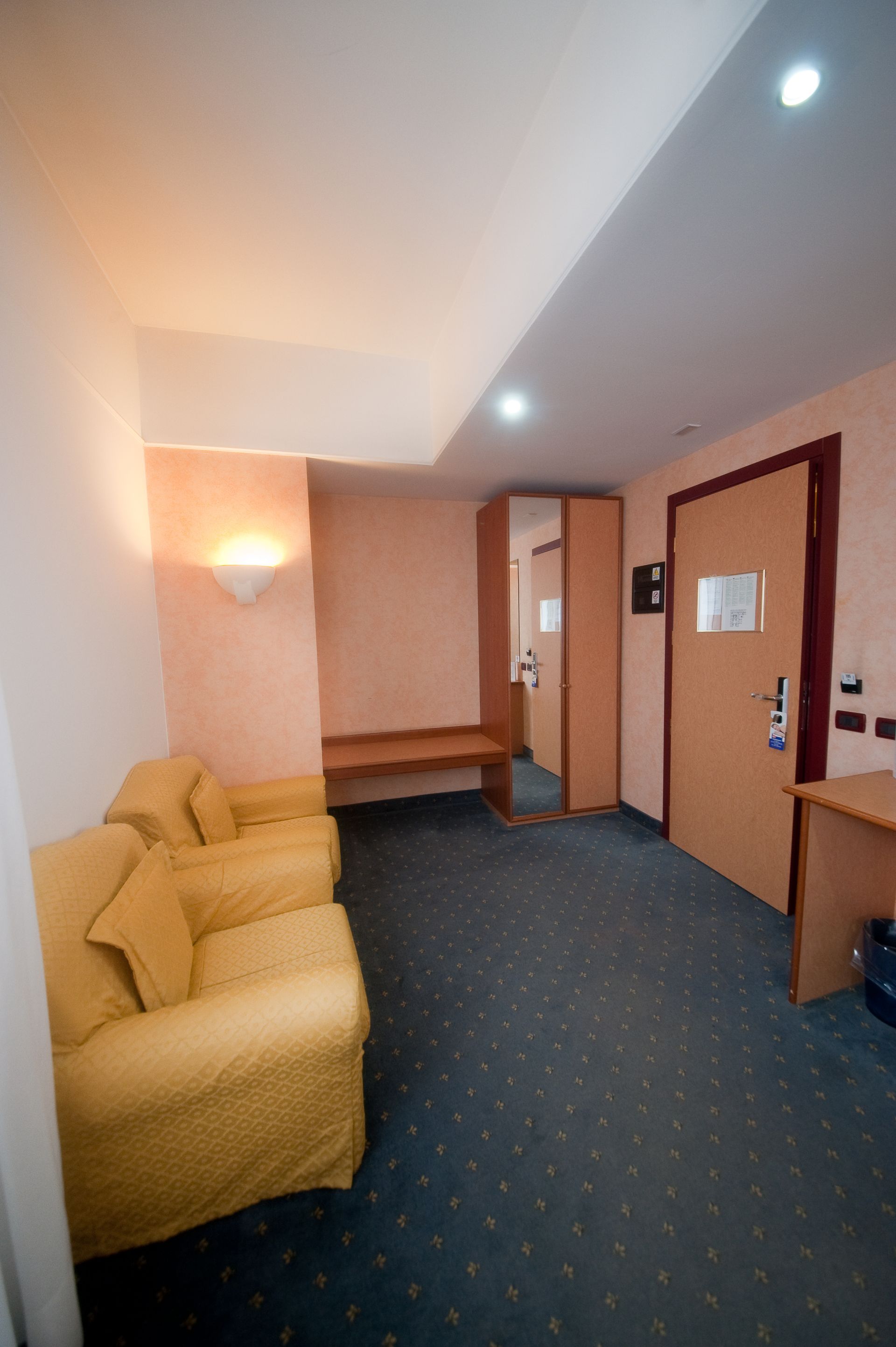 Interno di una camera d'albergo con divano giallo, moquette blu, porta marrone e pareti color pesca.