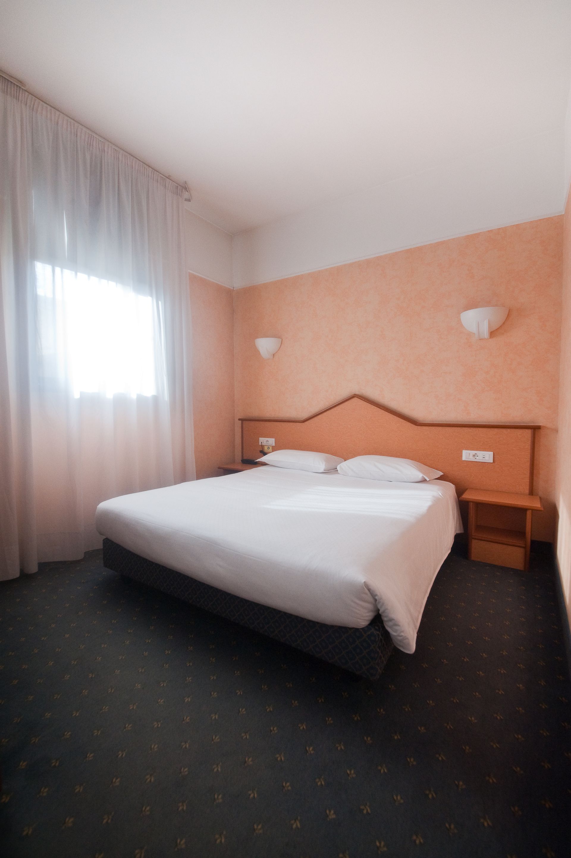 Camera da letto con letto, comodino e finestra. Parete arancione, biancheria da letto bianca e moquette blu.