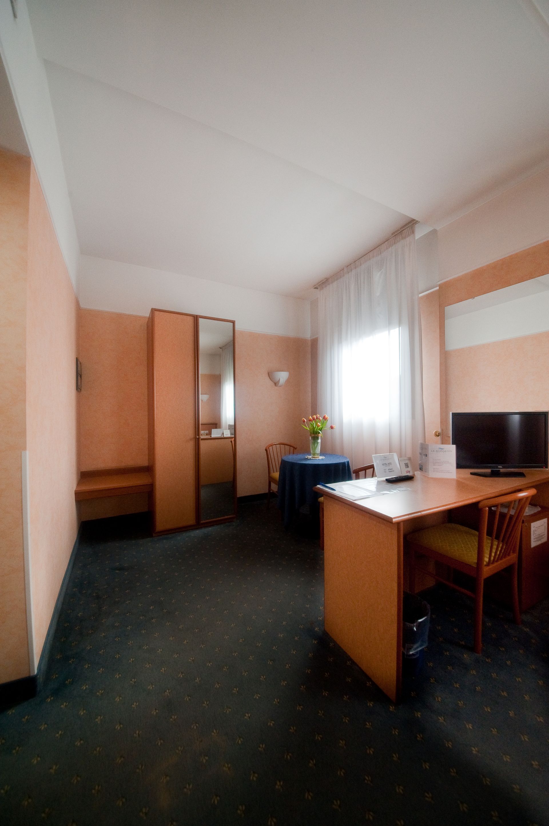 Camera d'albergo con scrivania, letto, armadio e tavolo con fiori.