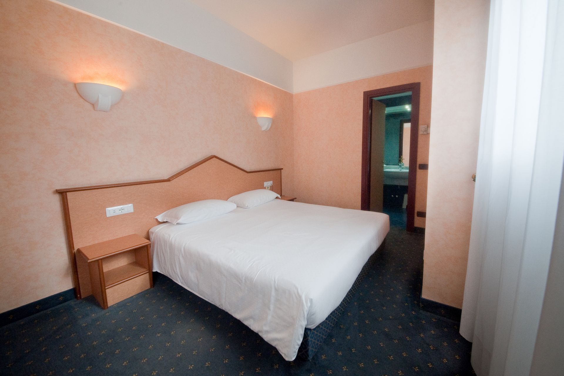 Camera d'albergo con letto, comodino e accesso al bagno. Pareti color pesca, moquette blu.