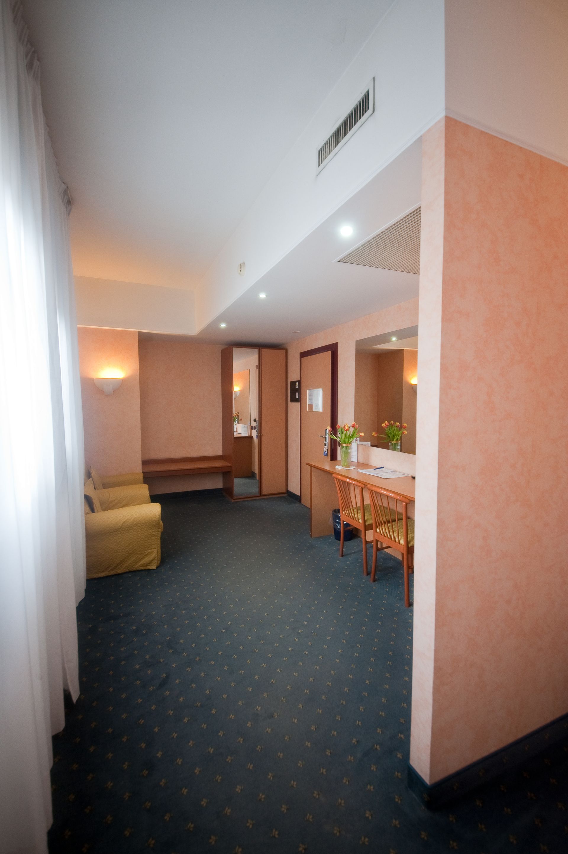 Interno della camera d'albergo: pareti beige, moquette blu, scrivania, divano, porta del bagno, tende bianche.