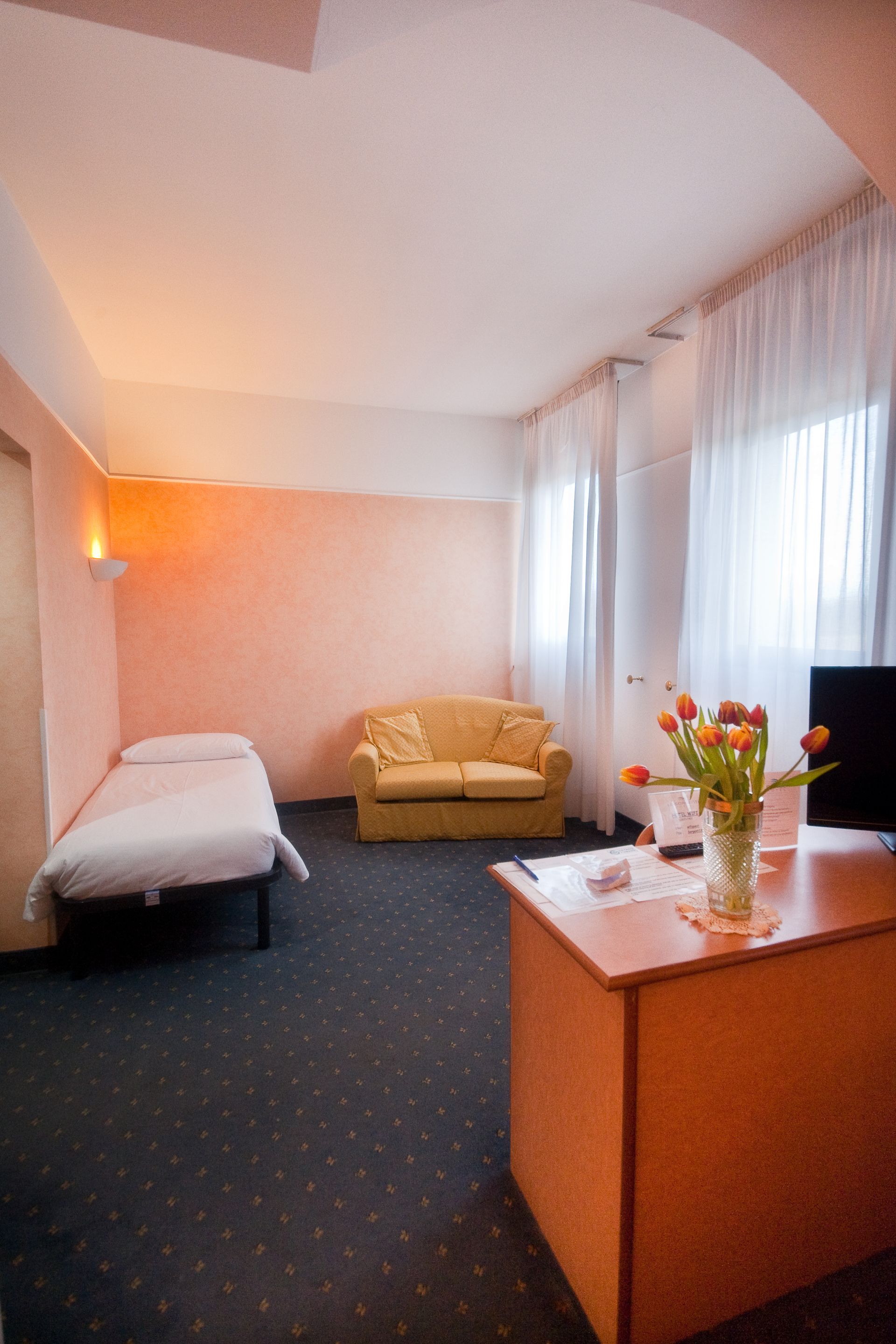 Camera d'albergo con letto, divano, scrivania con fiori e tende trasparenti.