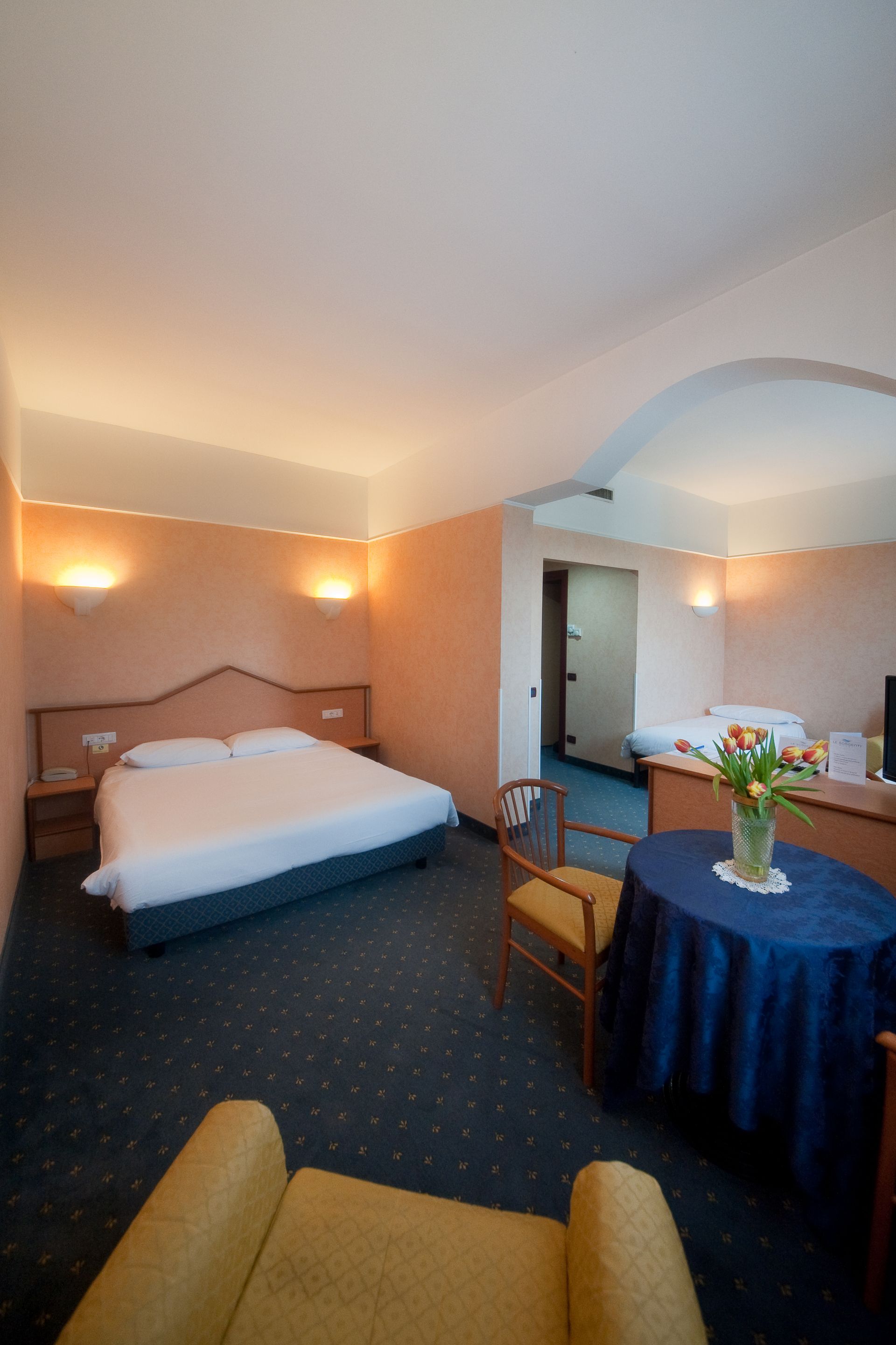 Camera d'albergo con letto, tavolo, sedie e un arco che conduce a un secondo letto.