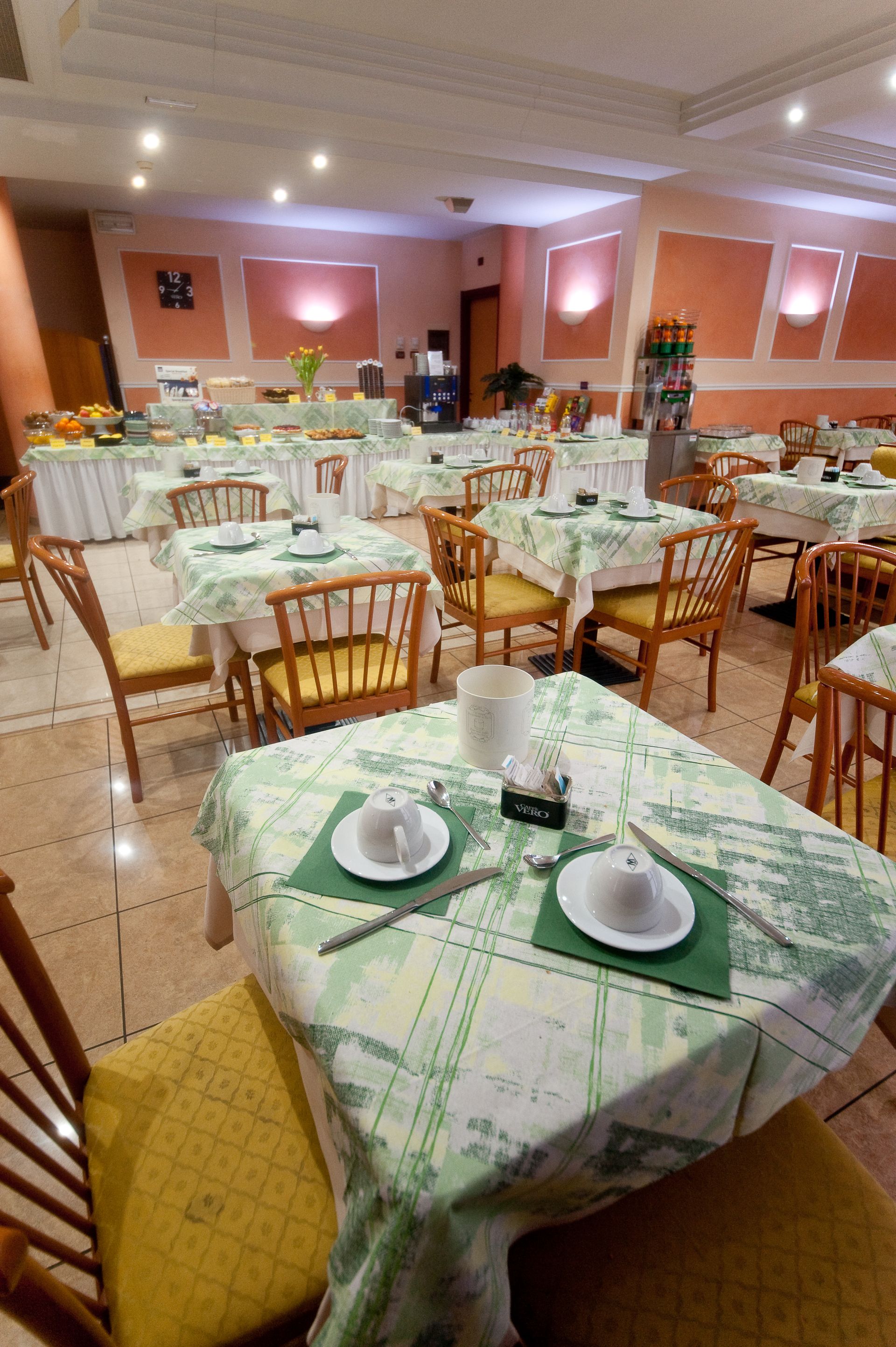 Sala colazioni dell'hotel con tavoli apparecchiati per il servizio, buffet sullo sfondo.