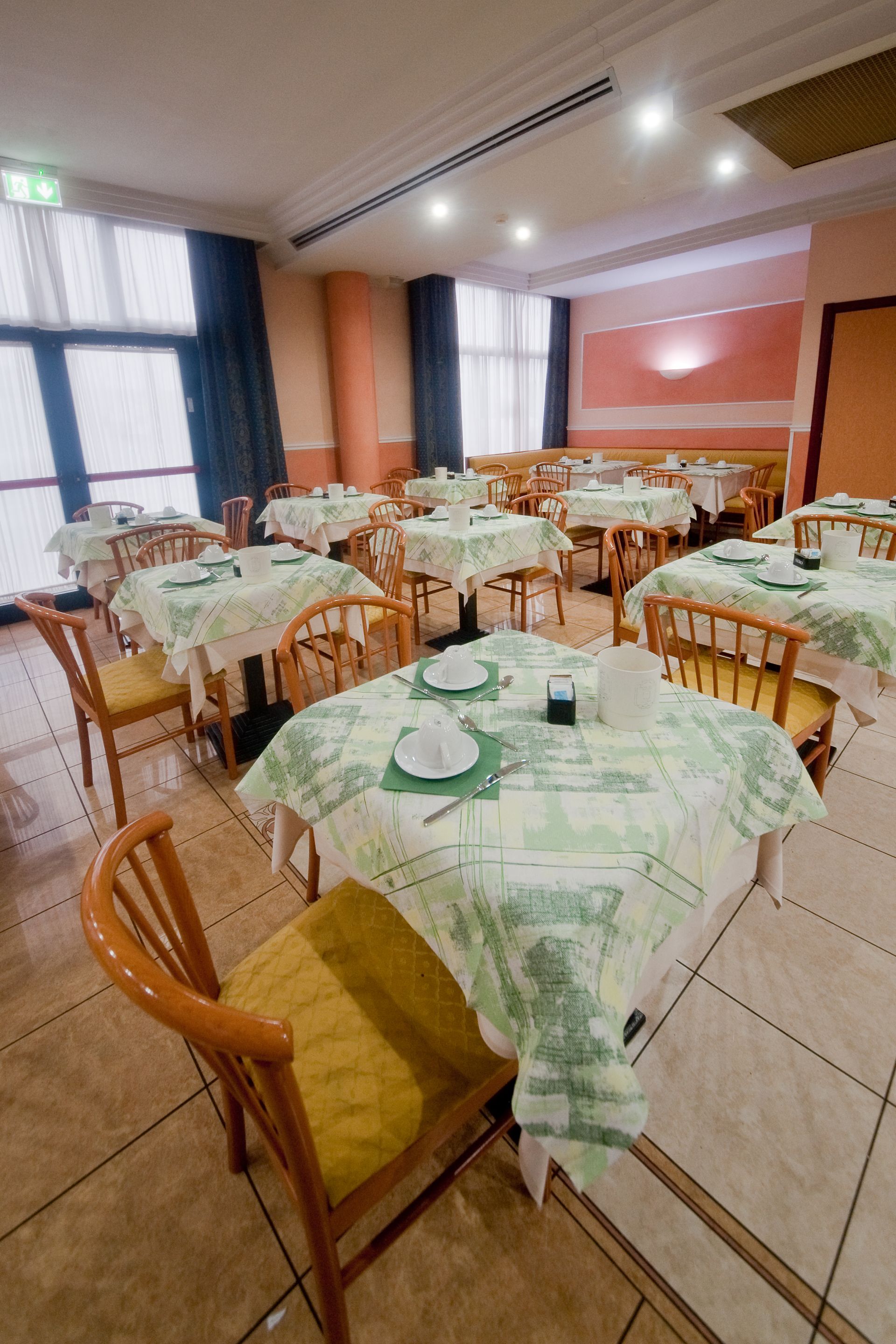 Sala da pranzo del ristorante con tavoli apparecchiati per il servizio, tovaglie a fantasia, sedie, finestre e pareti neutre.